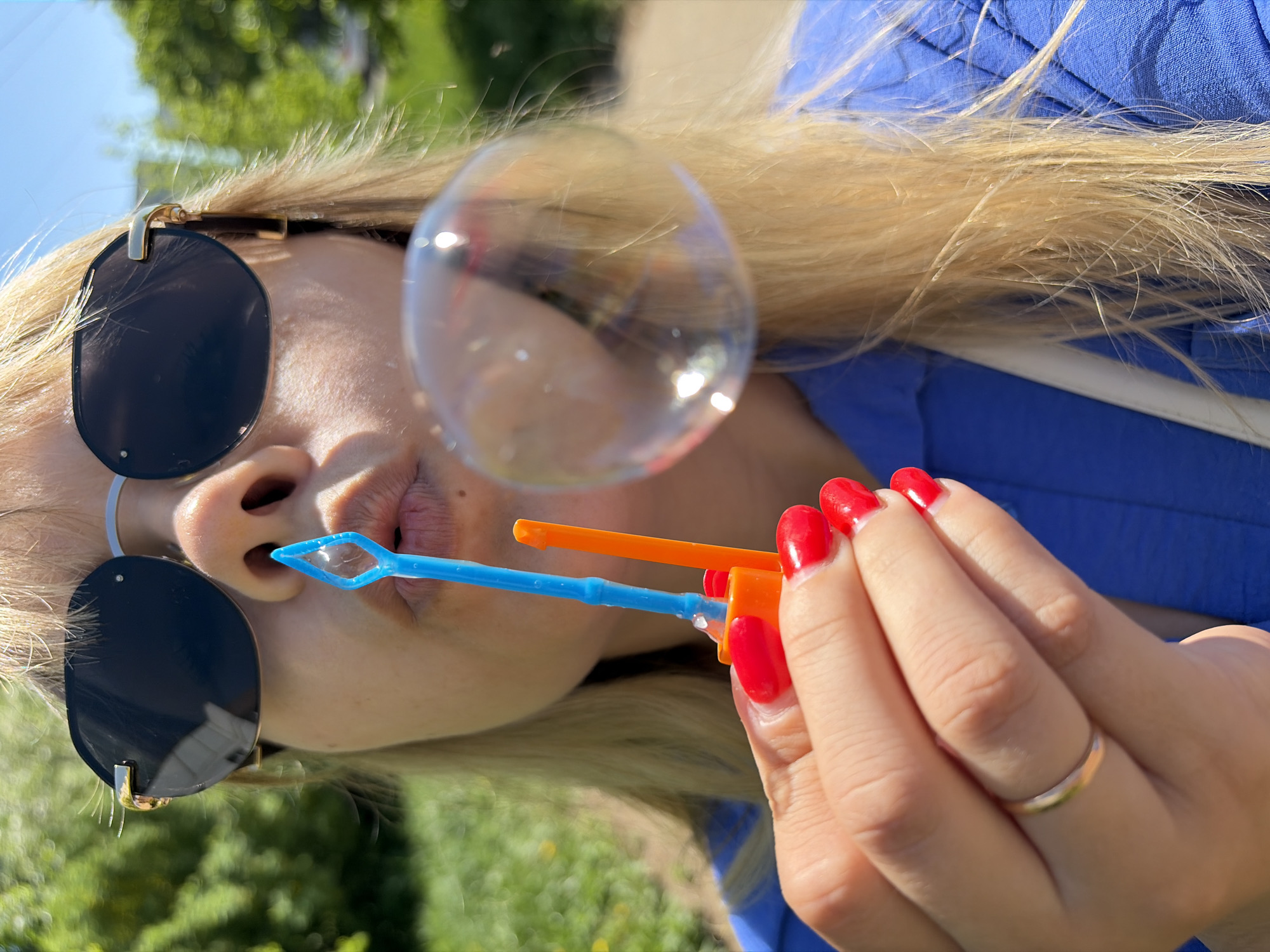 A woman with blonde hair, wearing dark sunglasses and a blue top, is blowing bubbles. She is holding a blue and orange bubble wand, and a bubble is floating near her face. Her fingernails are painted red, and she is wearing a gold ring. The photo appears to have been taken outdoors, possibly in a park or garden, with trees and sky visible in the background. The exact location is the city of Chaikovsky, Russia.