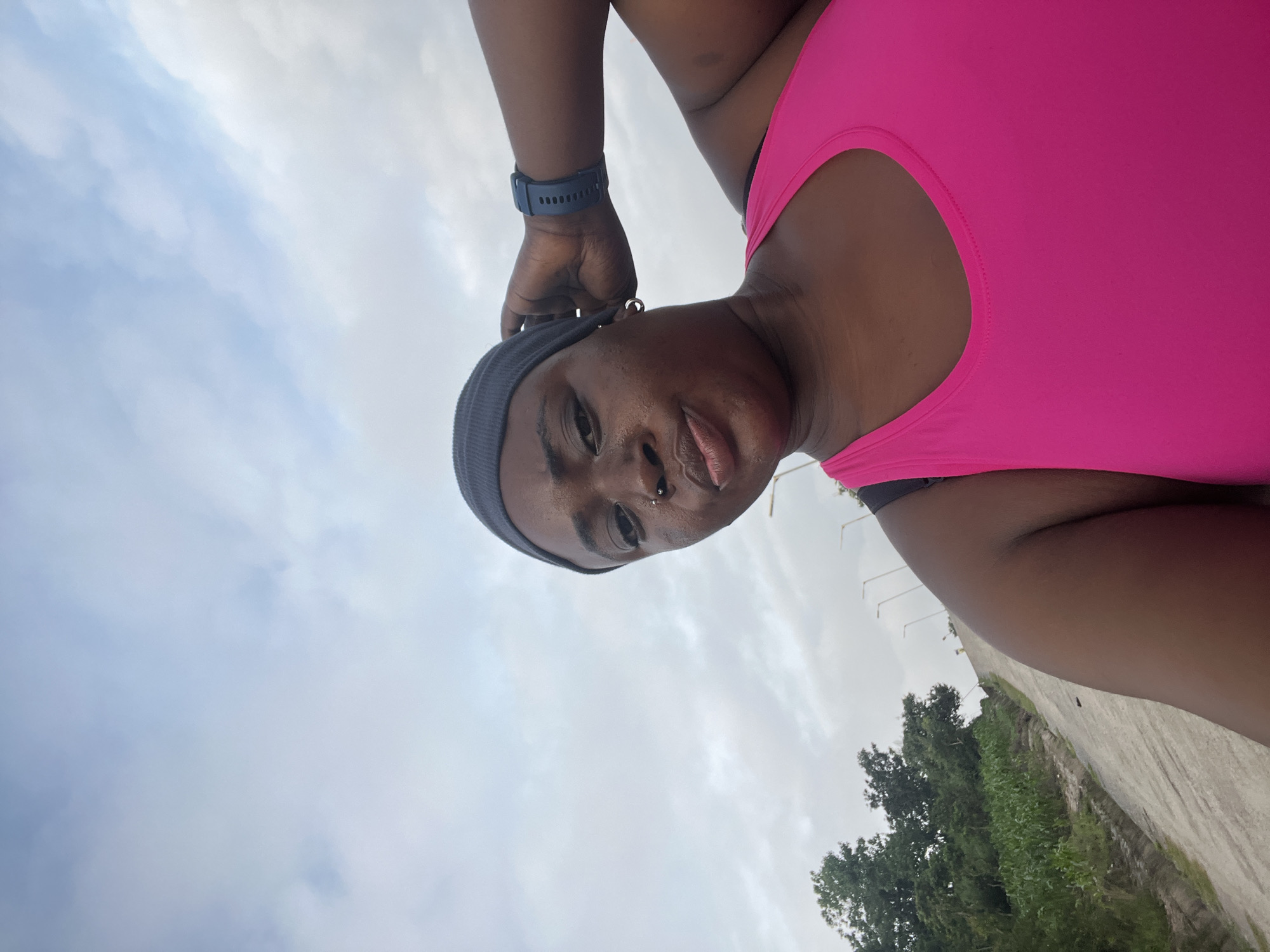 A medium shot shows a Black woman with dark skin and a round face. She is wearing a dark grey beanie, a bright pink athletic tank top, and a dark blue digital watch on her left wrist. She has a nose ring and a hoop earring in her left ear. Her right hand is raised and resting on her head. Her left arm is bent at the elbow. She is looking towards the camera with her mouth slightly open. The background is an overcast sky with light clouds. To the left, there is a paved road that slopes upwards, bordered by green vegetation and trees. Streetlights are visible in the distance.