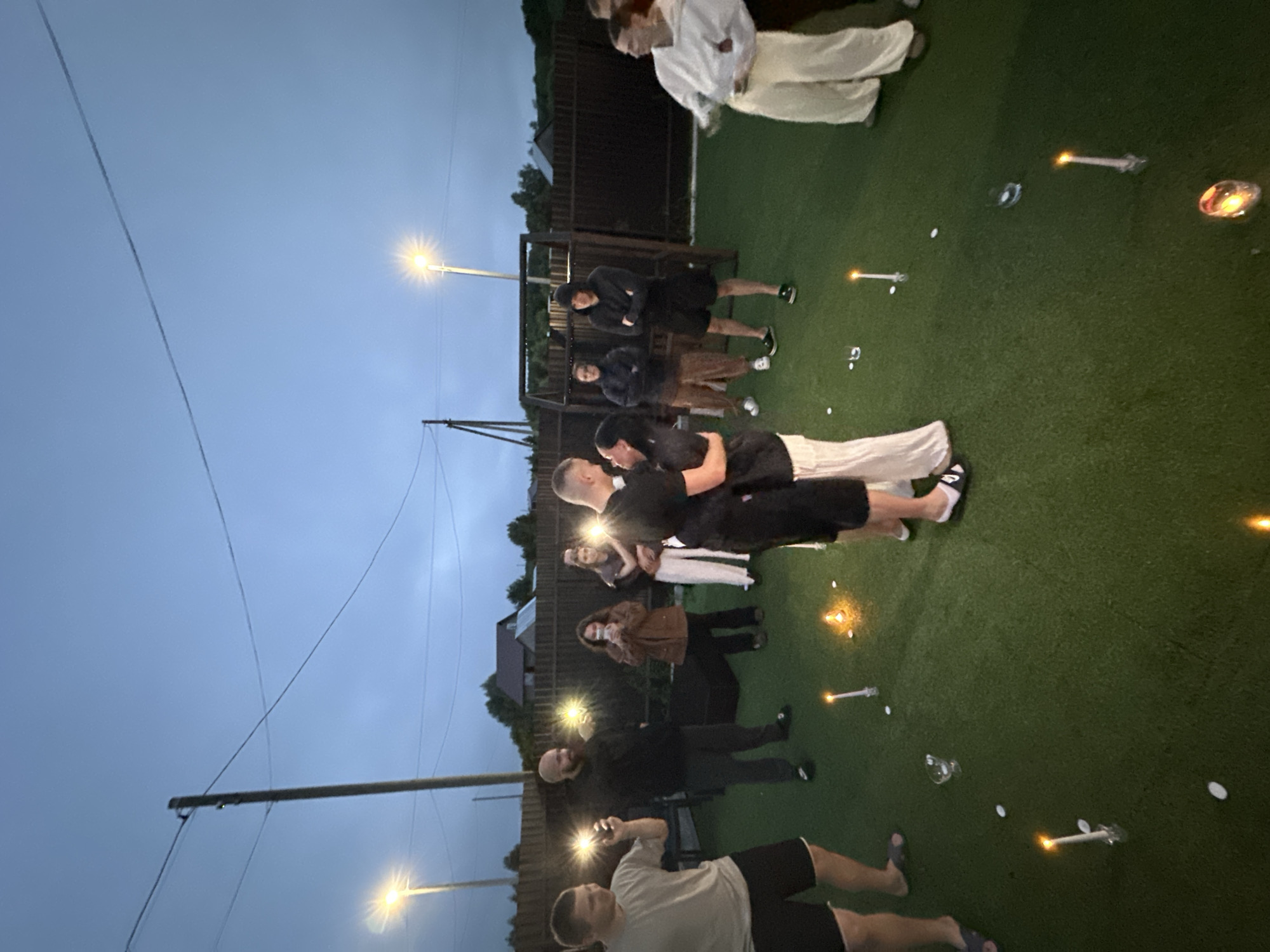 The image depicts an outdoor gathering at dusk in Karsha, Russia. A group of people are present, with a central focus on a couple in the foreground embracing and appearing to kiss. The couple is dressed in dark athletic wear, with one person wearing light-colored pants.
The setting appears to be a grassy area, possibly a backyard or park, with a fence in the background and some residential structures visible. Several candles are lit and scattered on the grass, creating a warm, atmospheric glow. A streetlamp with a bright light is also visible, illuminating the scene. The sky is overcast and a deep blue, indicating twilight.
Other individuals in the group are standing or observing, some holding what appear to be sparklers or small lights, adding to the celebratory or romantic ambiance. The overall mood seems intimate and joyful. The image is oriented vertically. There is no visible text in the image.
