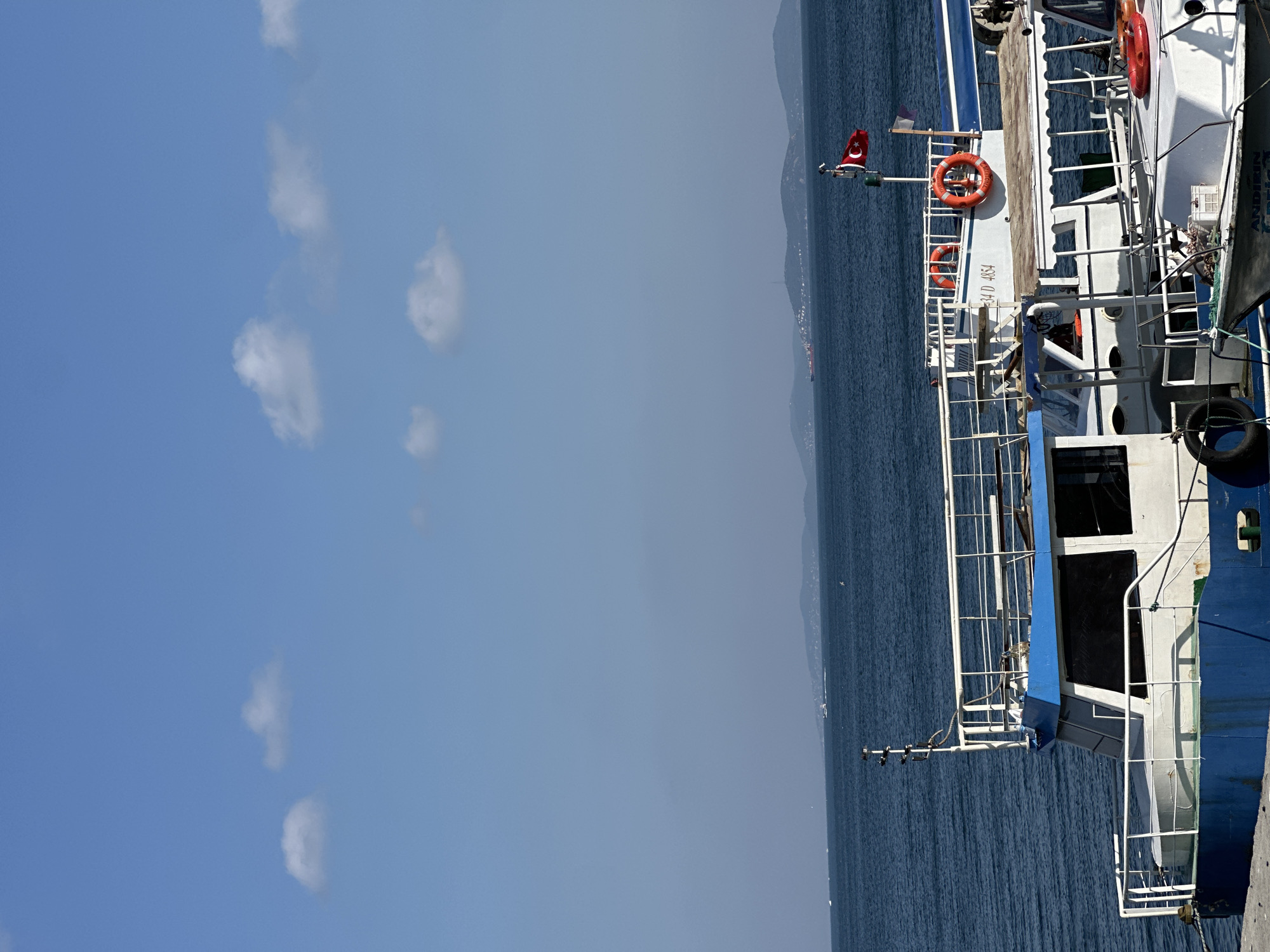 This is a photograph taken from a dock in Istanbul, Turkey, looking out over the sea. The sky is a clear blue with a few scattered white clouds. In the foreground, there are several boats docked. The closest boat has a blue and white hull, and the upper deck is white with blue accents. There are life preservers visible on the boats. In the middle ground, the deep blue sea stretches out towards a distant shoreline with rolling hills. A Turkish flag is flying from a pole on one of the boats. A larger ship can be seen on the horizon. The overall scene depicts a calm day at the harbor.