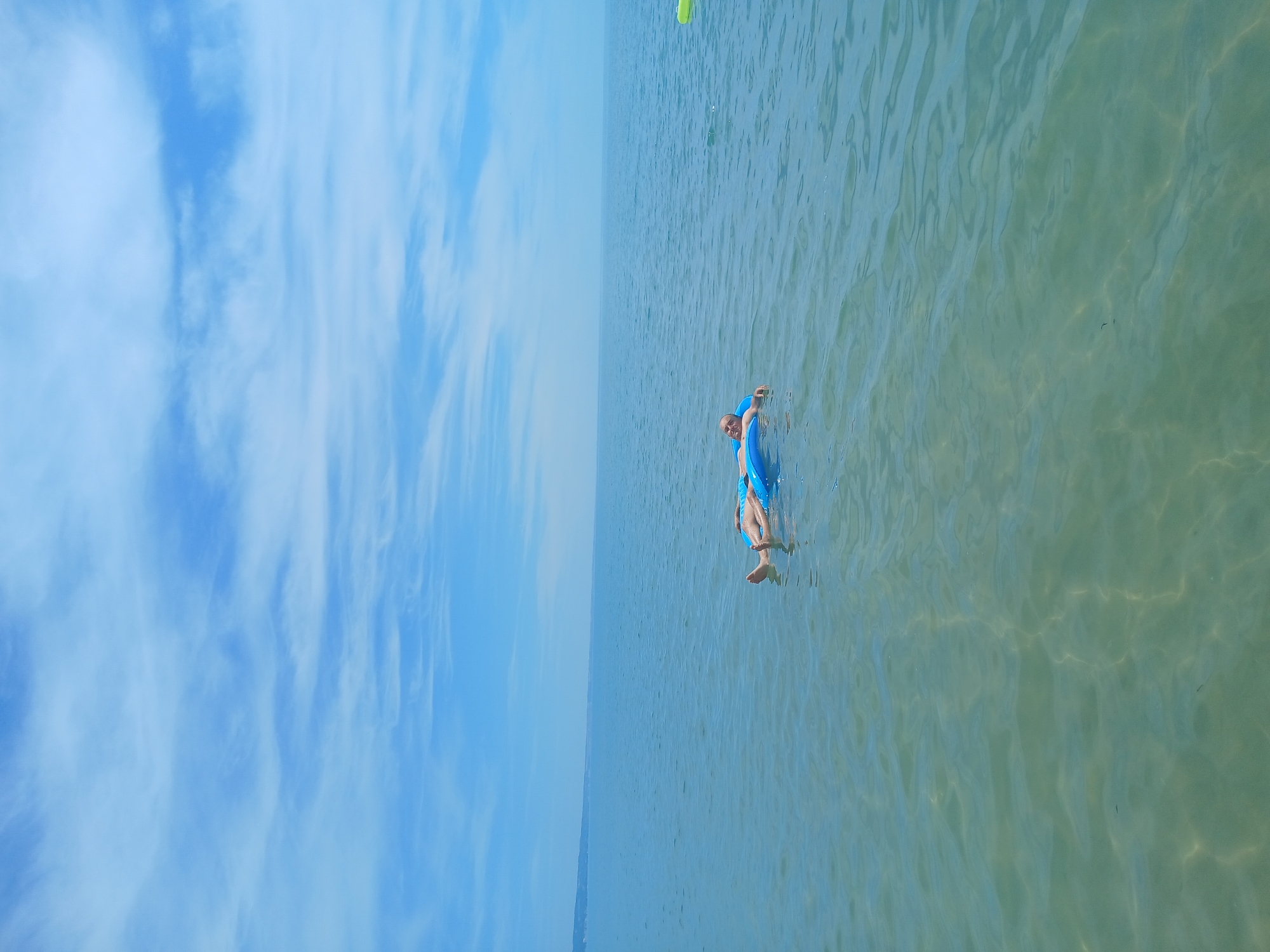 A person lies on a bright blue inflatable raft in the calm, shallow sea off the coast of Kranevo, Bulgaria. The water is clear and translucent, revealing the sandy seabed below. The sky is a brilliant blue, adorned with wispy white clouds. In the distance, a faint, hazy shoreline can be seen. The overall scene conveys a sense of peaceful relaxation on a sunny day.