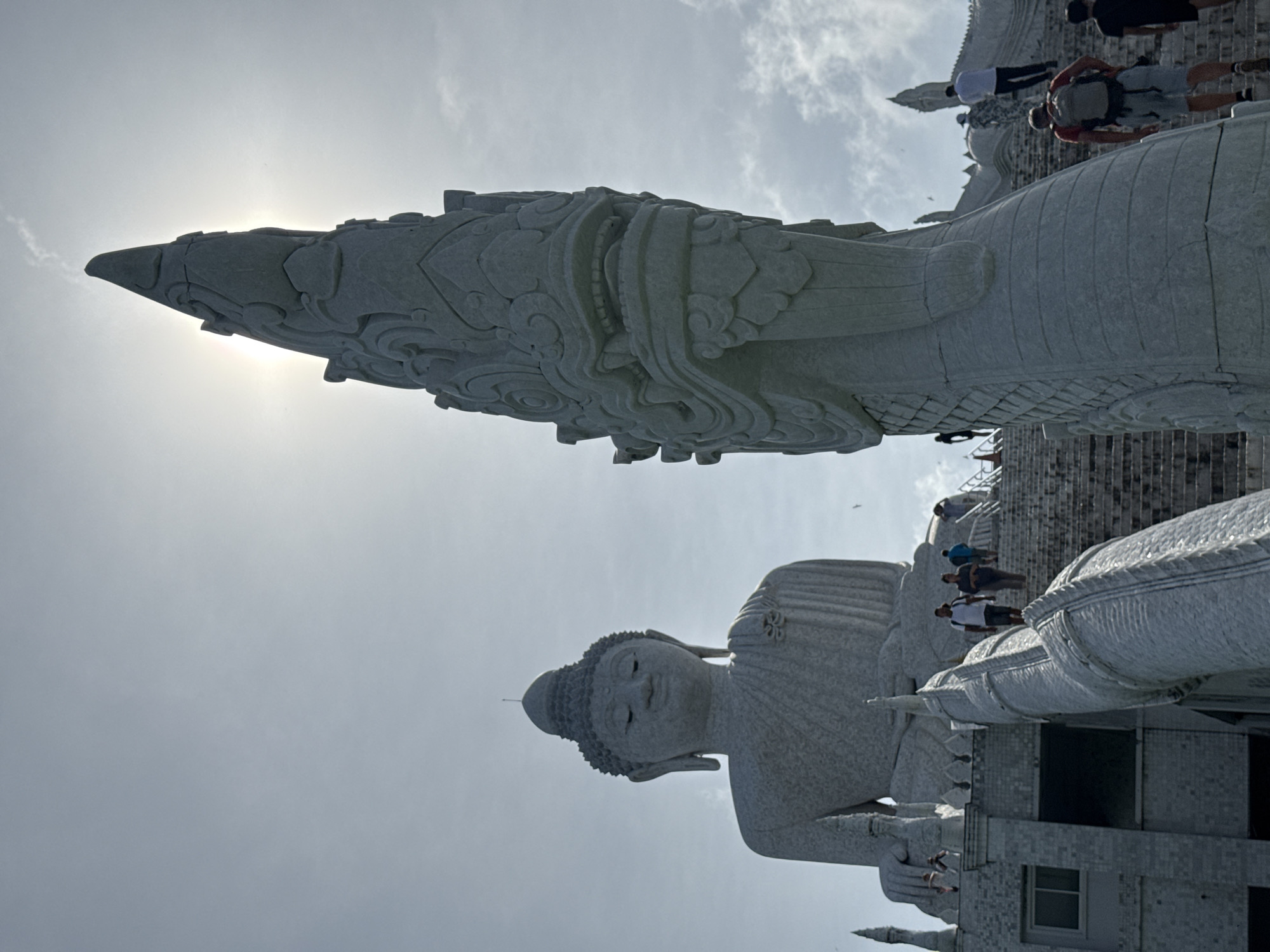 The image captures the Big Buddha of Phuket, a colossal statue situated atop Nakkerd Hills, near the city of Karon, Thailand. The viewpoint is from the base of the stairs leading up to the statue. In the foreground, a large, intricately carved stone serpent sculpture dominates the right side of the frame, its head partially obscured by the bright sunlight filtering through a cloudy sky. The serpent's scales and ornate features are clearly visible. To the left, the massive white marble statue of the Big Buddha is seen in the distance, seated serenely with its eyes closed. People are ascending the wide stone staircase, some are in groups, while others walk individually, their figures silhouetted against the bright sky. The environment appears to be outdoors, with a bright, overcast sky. The overall impression is one of grandeur and spiritual significance.