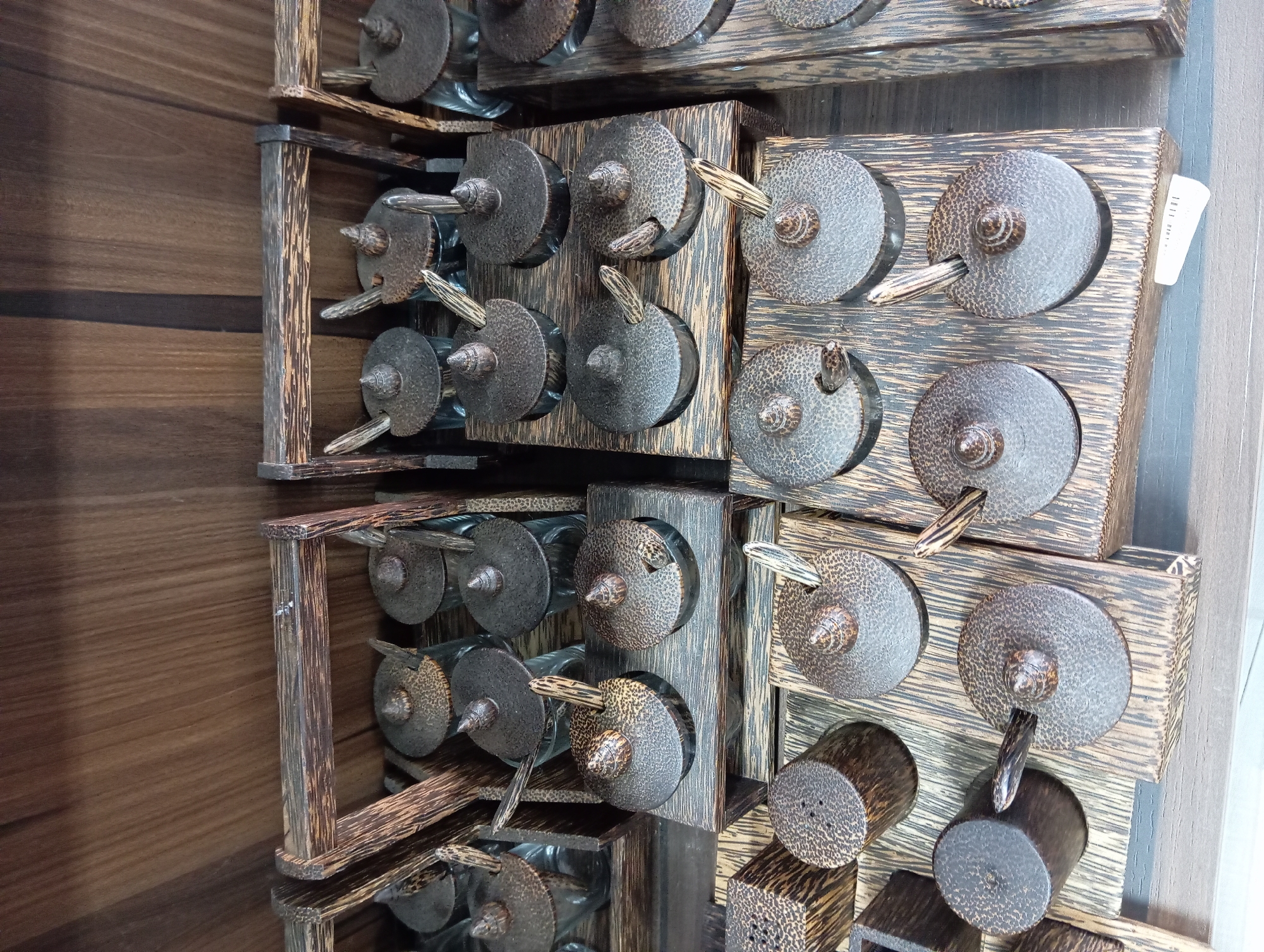 The image shows a close-up view of several wooden spice containers with lids and small spoons, arranged in wooden trays. The containers have a dark, textured pattern, likely from the wood itself, and a conical finial on each lid. The background appears to be a wooden shelf or wall. The arrangement suggests a retail display, possibly in a souvenir shop or market. This scene could be found in Nha Trang, Vietnam, a popular tourist destination known for its beaches and local markets where such handcrafted items are often sold.