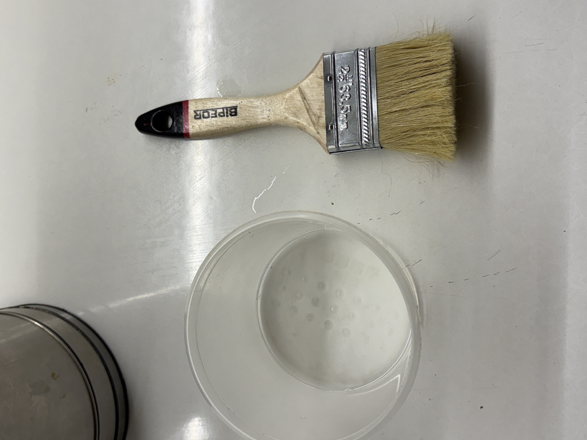 A top-down shot depicts a paint brush with a wooden handle and natural bristles lying horizontally on a white surface. Beside the brush, a clear plastic container holds a milky white liquid with visible globules, possibly paint thinner. In the lower left corner, part of a metallic paint can is visible. No people are present in the image. The location cannot be determined to be in Yerevan, Armenia, or any other specific location.