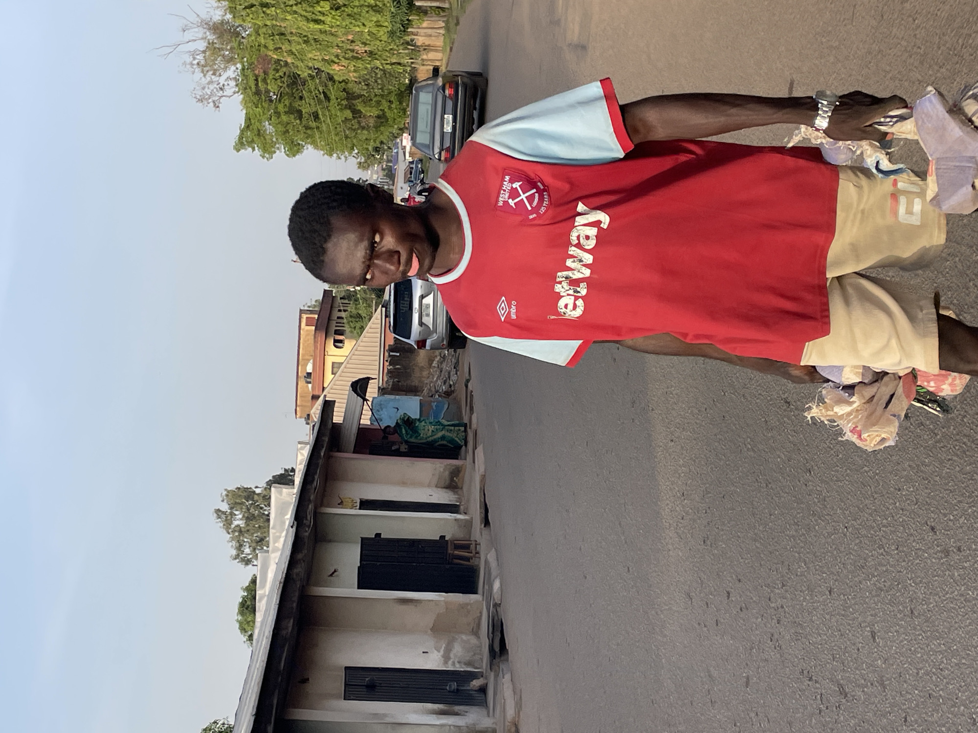 A dark-skinned man stands on a paved road, looking directly at the camera with a playful expression, his tongue slightly out. He is wearing a red and light blue short-sleeved football jersey, prominently featuring the "WEST HAM UNITED" club crest with "125 YEARS" underneath, the "Betway" sponsor logo across the chest, and the "UMBRO" sportswear logo. He also wears light-colored shorts with a "FILA" logo visible on the left leg. A metallic watch is visible on his left wrist. In his right hand, he carries a bundle of multi-colored clothes or rags.
The setting is an outdoor street scene, likely an urban or semi-urban area in Jos, Nigeria. In the background, other vehicles, including a black sedan and a silver car, are visible on the road. To the left, several one-story buildings with corrugated roofs and what appear to be shuttered windows line the street. One building shows a person lying down, partially covered by a green cloth, on an elevated porch area. Lush green trees are scattered above the buildings and in the distance. The sky is clear and light blue, indicating daytime, possibly late afternoon given the soft light.