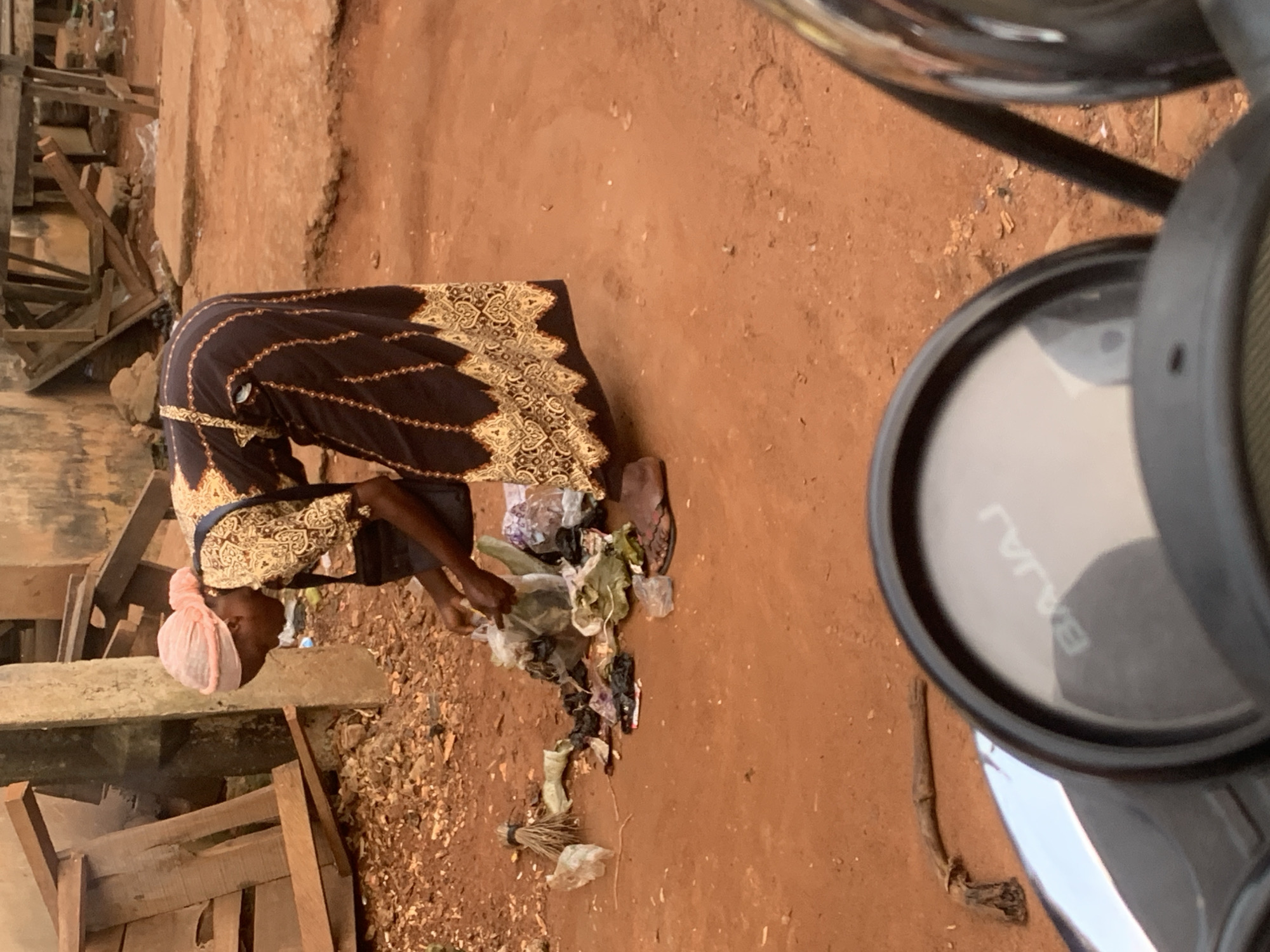 A woman wearing a pink headscarf and a dark dress with gold trim bends down to collect trash in a dirt lot in Shagamu, Nigeria. Scattered on the ground are various pieces of garbage, including plastic bags, wrappers, and other debris. A broom lies on the ground to the left of the woman. In the foreground, a motorcycle's dashboard is visible, with the word "BAJAJ" clearly legible on the speedometer. The background shows a rustic building and wooden structures.