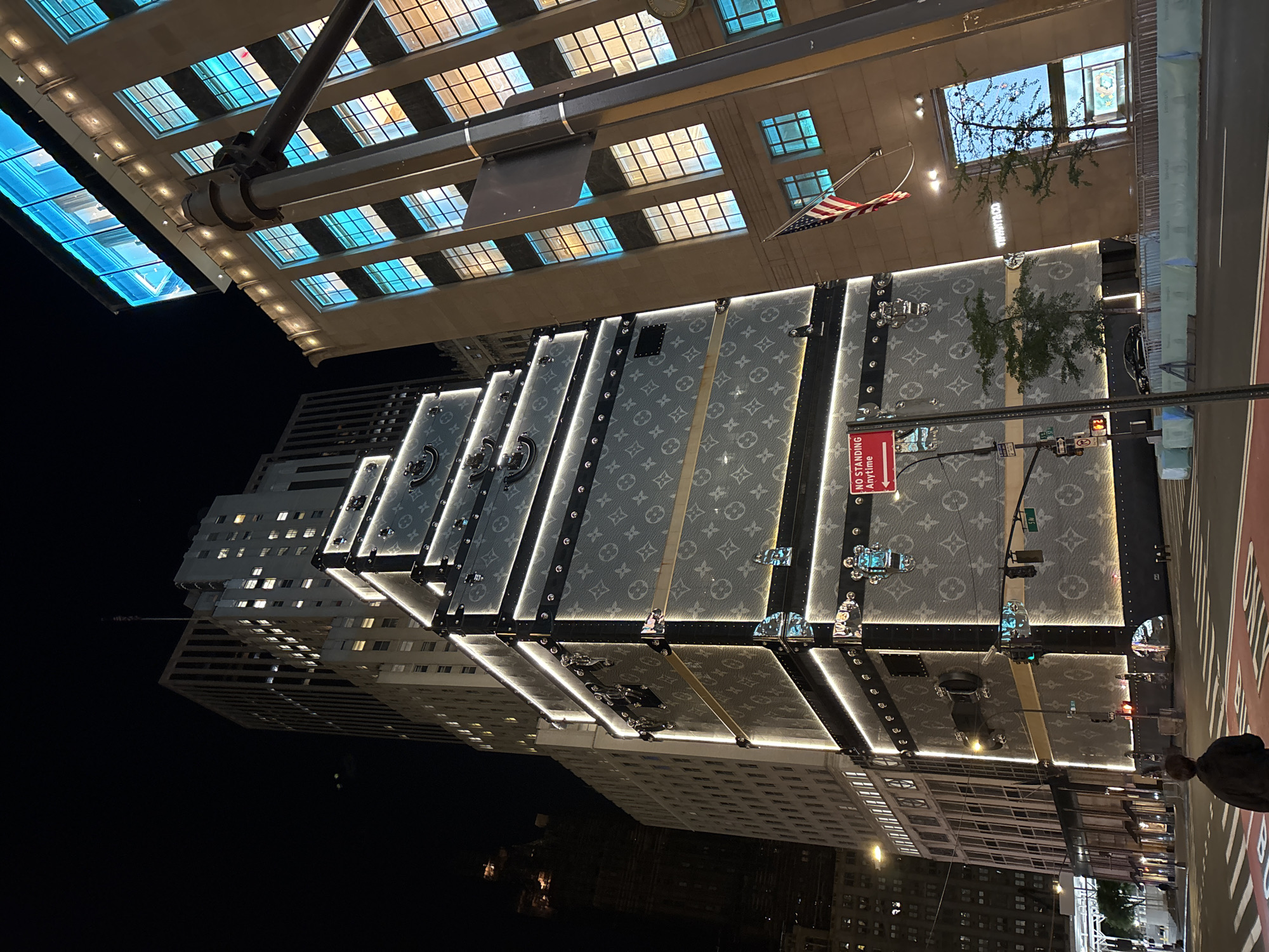 An urban night scene captures a brightly illuminated building facade designed to resemble giant Louis Vuitton trunks, dominating the right side of the frame. The facade features the iconic LV monogram pattern and is outlined with brilliant white LED strips, creating a glowing effect against the dark sky. The "trunks" are adorned with faux straps, buckles, and hardware, mimicking the luxury brand's luggage.
Above this striking building, an elevated structure extends across the upper part of the image. This structure has a beige underside with rows of grid-like windows, some emitting a cool blue light, while others glow with a warmer, amber hue. A heavy dark metal beam runs prominently beneath this overhead structure. An American flag hangs from a pole near the center of the image, fluttering slightly.
In the foreground, a city street with red road markings is visible, along with a curb and a traffic light showing a red signal. A green street sign indicates "36th St." A red rectangular sign reads "NO STANDING ANYTIME." A person is partially visible on the far right, near the street. In the background, other tall, dark city buildings with illuminated windows can be seen, reflecting the bright lights of the Louis Vuitton facade. The time of day is clearly night, evidenced by the deep black sky and the reliance on artificial light sources, creating a dramatic and vibrant city atmosphere.