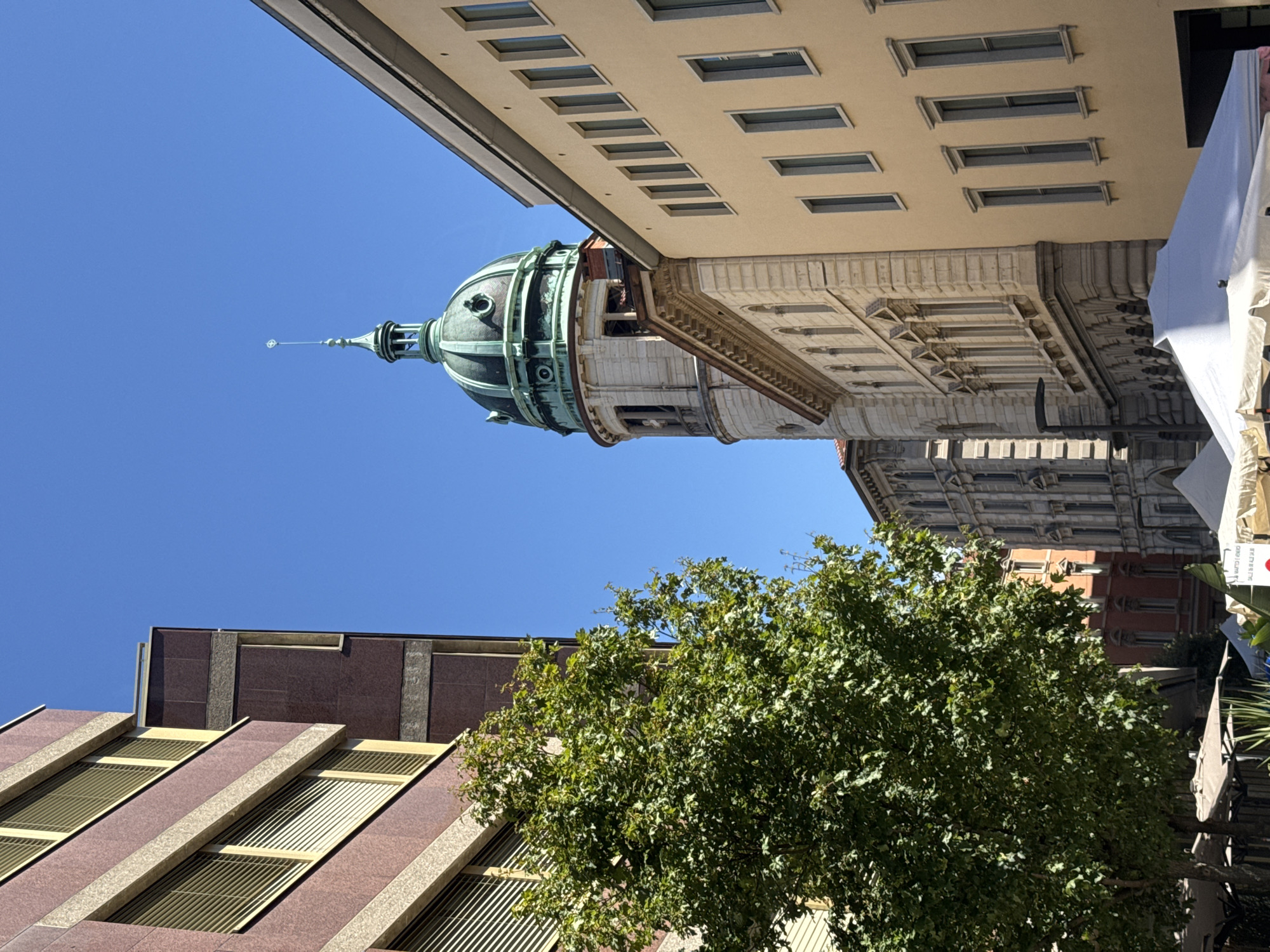 The image depicts a street scene in Lugano, Switzerland, under a clear blue sky. In the foreground, a large, leafy green tree partially obscures the view of adjacent buildings. To the left, a modern building with a reddish-brown facade and horizontal window blinds is visible. To the right, a pale yellow building with a series of tall, narrow windows lines the street.
Dominating the middle ground is a prominent building with a large, green dome topped with a spire. This structure, likely a religious or civic building, features a light-colored stone tower with arched windows. Adjacent to it, another building with a light stone facade and classical architectural elements, including arched windows and decorative molding, is visible. In the lower portion of the image, white tents or canopies are set up, suggesting an outdoor market or event. The overall impression is of a sunny day in a European city, with a mix of architectural styles.