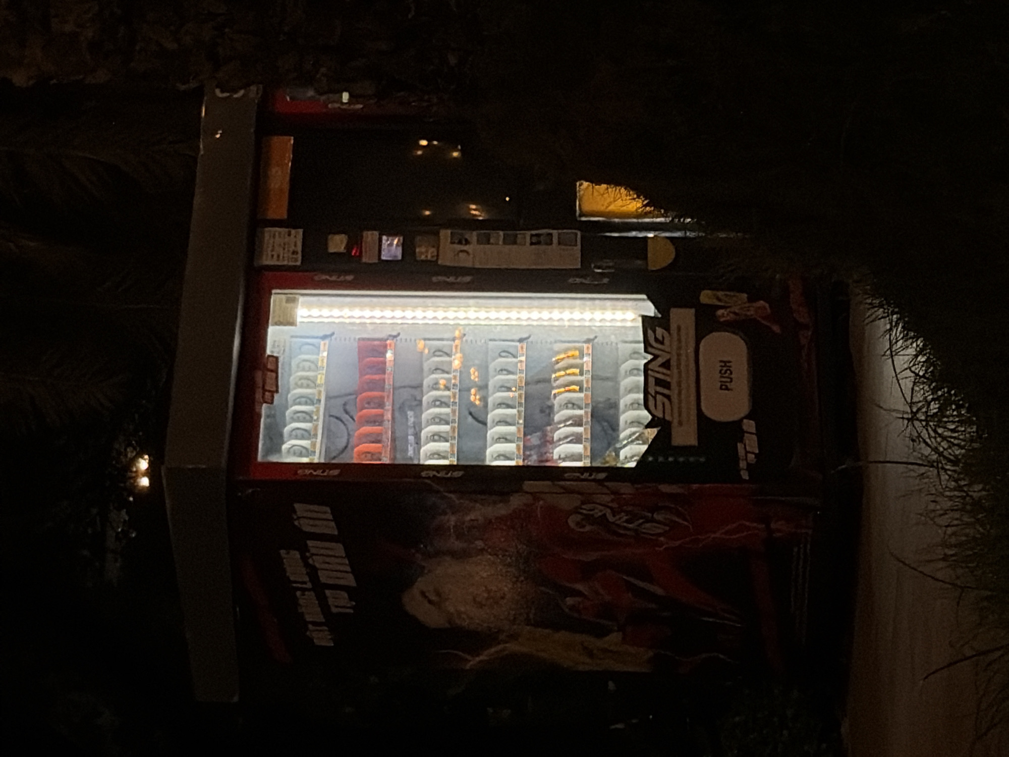 This nighttime image depicts a "Sting" branded vending machine illuminated internally, standing outdoors in Thủ Đức, Vietnam. The vending machine features a red and black exterior with a large side graphic of a person and the Vietnamese text "MỘT NĂNG LƯỢNG TẬP ĐẦU ĐỜN" (translating to "ONE ENERGY FIRST GROUP/BEGINNING"). Inside the glass front, multiple rows of beverage cans are visible, appearing in white/silver, orange/red, and yellow/gold colors. A "Sting" logo and a prominent "PUSH" button are located below the display window. To the right, various informational stickers and a payment/dispensing area with a warm yellow light reflection are observed. The machine rests on a paved surface, with dark foliage and grass visible in the foreground and surrounding background, indicating an outdoor, unpopulated setting.