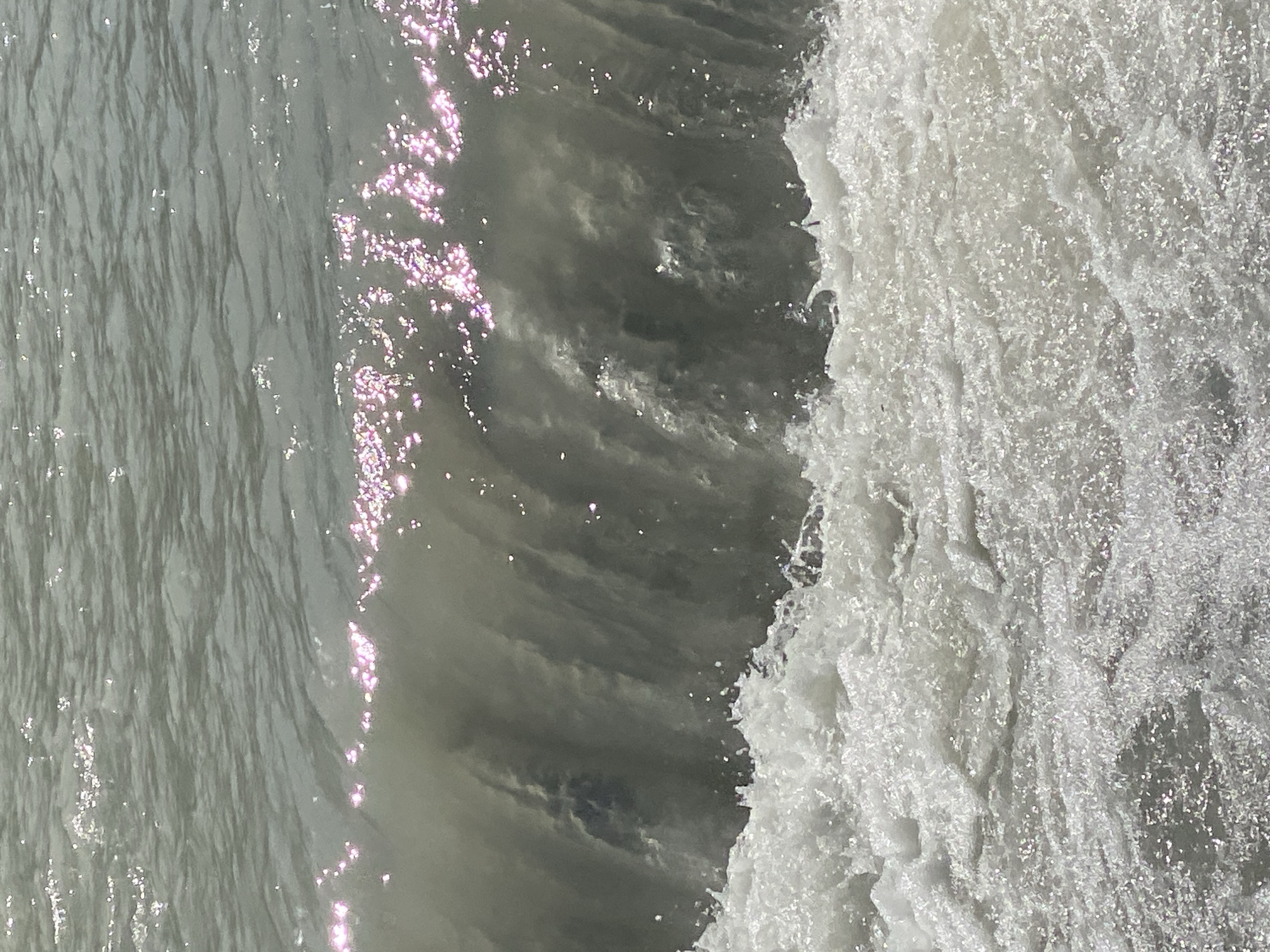 The image shows a close-up view of a breaking wave on a body of water. The water is a murky gray-green color, and the wave is churning with white foam at its base. Sunlight glints off the surface of the water, creating bright highlights. The background shows further ripples on the water. The scene is natural and appears to be a coastal or lakeside environment. There is no indication of urban setting or any landmarks that would identify it as being in Bishkek City, Kyrgyzstan.