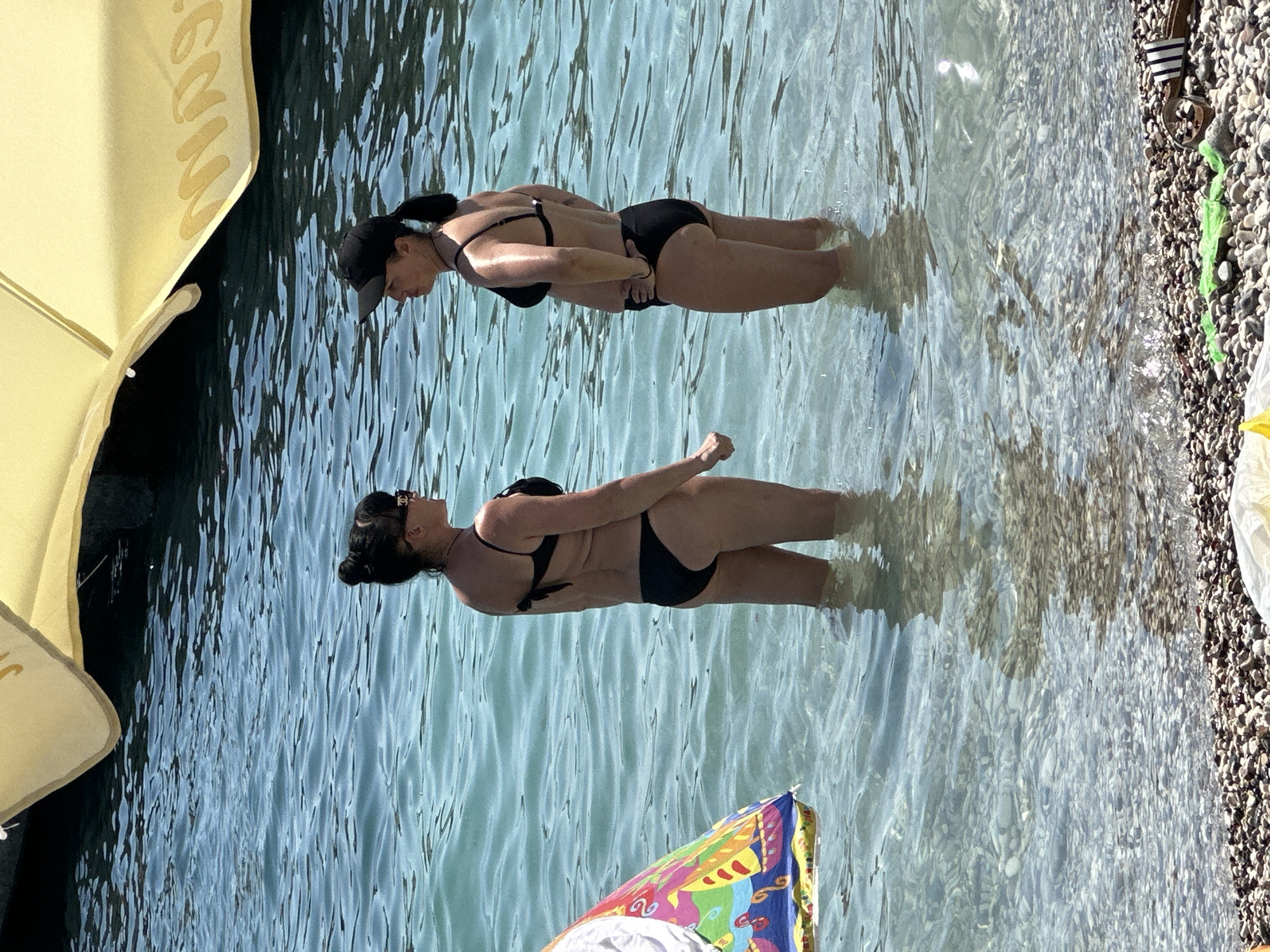 Two women stand in shallow, clear blue water at a pebble beach in Budva, Montenegro. The woman on the left has dark hair pulled into a bun and wears a black bikini. She is facing the woman on the right, who has dark hair tied back in a ponytail and wears a black bikini and a black baseball cap. She is looking at the woman on the left. To the left of the women is a colorful inflatable object, partially submerged. In the background, a yellow beach umbrella is visible, casting a shadow on the water. The sun is shining, creating ripples on the water's surface and reflecting light. The beach is composed of small, smooth pebbles.