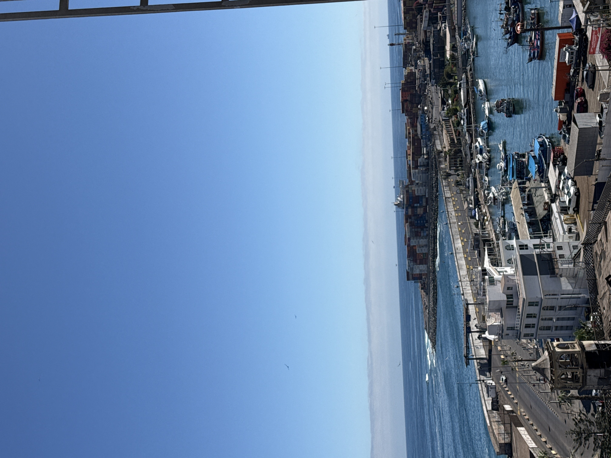 The image depicts a panoramic view of the port of Iquique, Chile. The clear blue sky stretches across the upper half of the frame, with a few birds soaring in the distance. The lower half of the image is dominated by the bustling port, featuring numerous cargo containers stacked high on the docks. Several ships, including a large cargo vessel and many smaller boats, are visible in the harbor. The waterfront is lined with industrial buildings and infrastructure, indicative of a busy commercial port. A road runs alongside the water, with a few vehicles present. In the foreground, there are architectural elements that suggest older buildings or structures, contrasting with the modern port facilities. The scene captures the dynamic activity of the port against a backdrop of the expansive ocean.