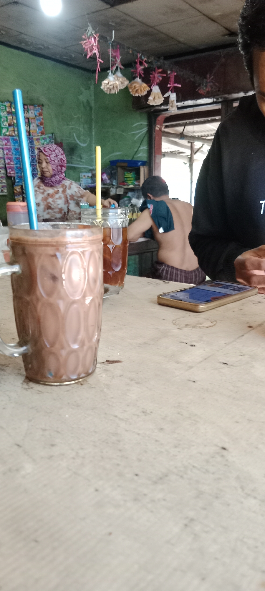 This is a medium shot, eye-level, indoor photograph taken in the city of Ngargosari, Indonesia. The scene appears to be inside a small shop or eatery. In the foreground, a large glass mug filled with a brown beverage, likely chocolate milk, is on the left. A blue straw extends from the mug. To the right of the mug, a second glass filled with iced tea and a yellow straw sits on the same wooden table.
In the background, a woman wearing a pink hijab and a floral-patterned shirt is visible behind the counter, appearing to be working or serving customers. A man, shirtless and wearing striped shorts, is seated behind the counter, with a dark cloth draped over his shoulders. He is facing away from the camera and appears to be engaged in some activity.
Above the counter, decorative items, possibly related to a local celebration or festival, are suspended from the ceiling. These include red tassels and small sachets of what appear to be snacks or treats. The walls are painted a light green, and there are shelves stocked with various items behind the counter.
To the right of the frame, a hand holding a smartphone is visible, with the phone's screen displaying what looks like a social media feed or web page. The overall lighting suggests it is daytime, with a bright light source visible near the ceiling on the left. The table surface is made of light-colored wood with visible grain and some scuff marks. The atmosphere suggests a casual, everyday setting.