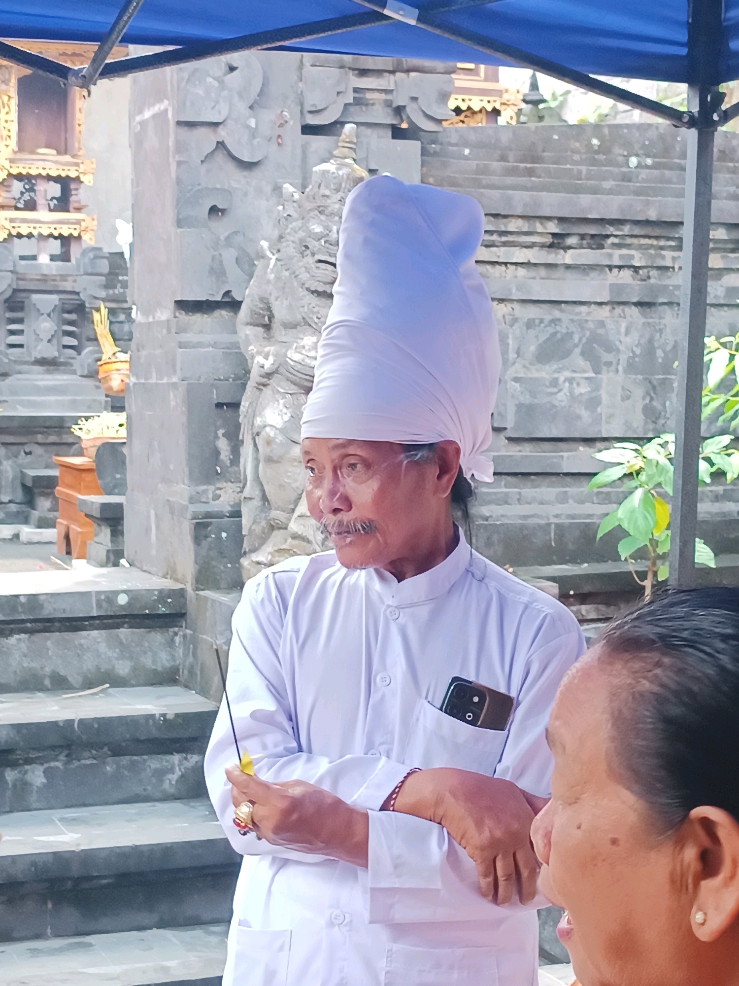A medium shot shows a man in traditional white clothing and a large white head covering, standing outdoors. He is holding a lit incense stick in his right hand and a yellow object in his left. A smartphone is visible in his chest pocket. To the right of the man, the side of another person's face is visible, their left ear with an earring is also visible. The background features stone steps, an ornate structure with gold accents, and a stone sculpture of a mythical creature. The scene appears to be at a religious or cultural site.