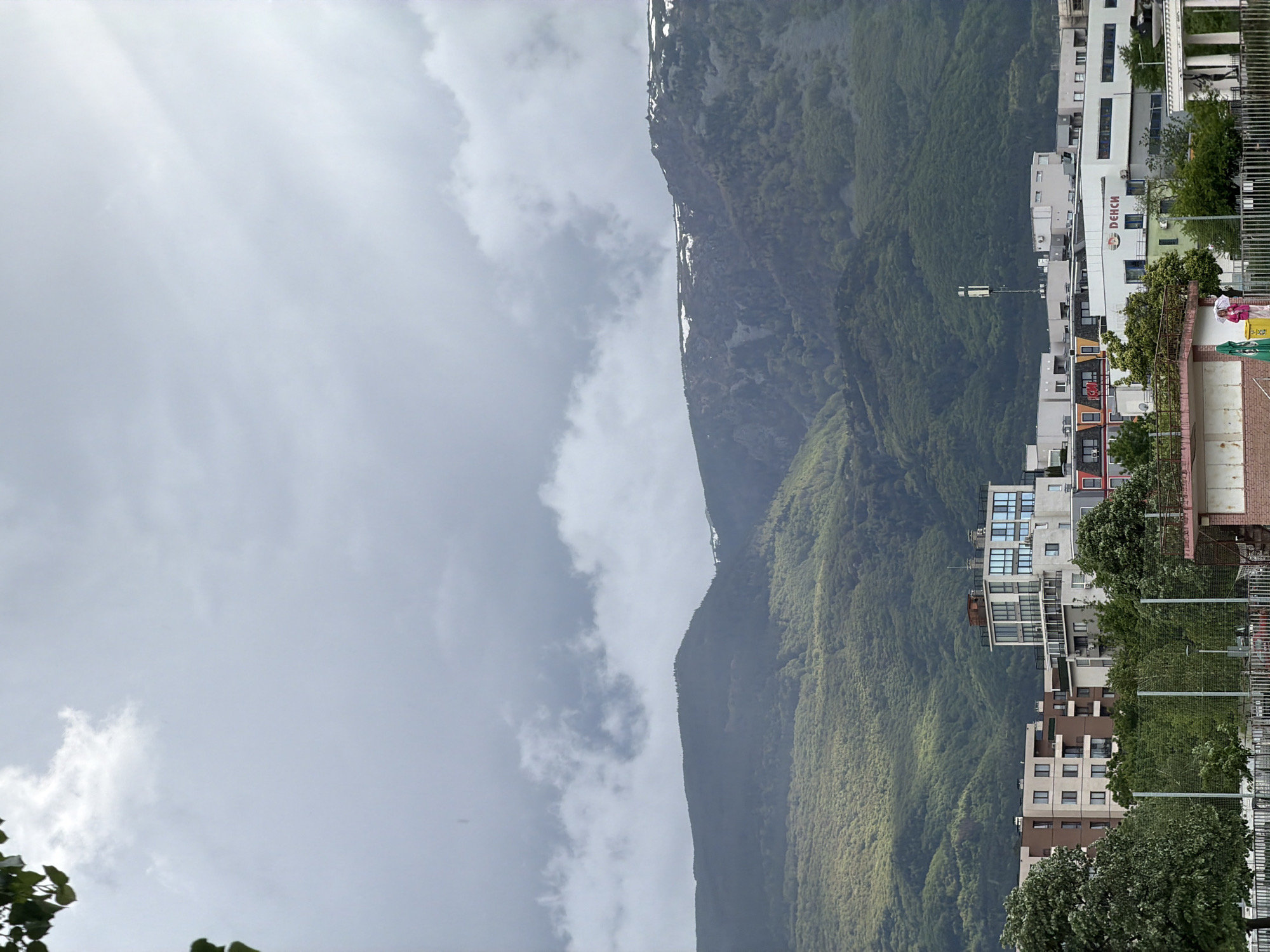The image shows a cityscape with mountains in the background under an overcast sky. On the right side of the image are several multi-story buildings with a mix of residential and commercial spaces. The mountains are covered in green vegetation. There is a billboard on one of the buildings that reads "DEKCHI." The location appears to be Sofia, Bulgaria, given the architectural style and landscape.