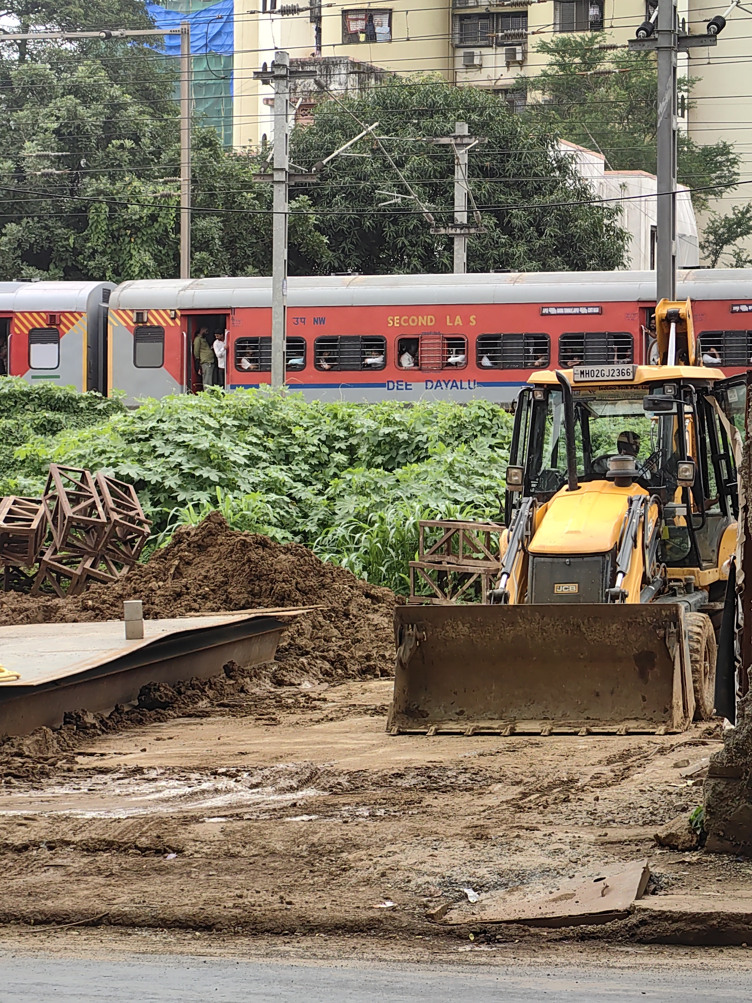 The image depicts a construction site in Mumbai, India, adjacent to a railway track. A yellow JCB backhoe loader is prominently positioned in the foreground, with its bucket resting on muddy ground. Behind the backhoe, a red passenger train is visible, with its doors open and passengers inside. The train appears to be stationary. The environment is characterized by dense green foliage bordering the tracks and a pile of dirt or soil next to the backhoe. The ground is uneven and muddy, indicating recent rainfall or ongoing construction work. The overall scene suggests an active construction or maintenance project taking place near a busy railway line within the urban landscape of Mumbai.