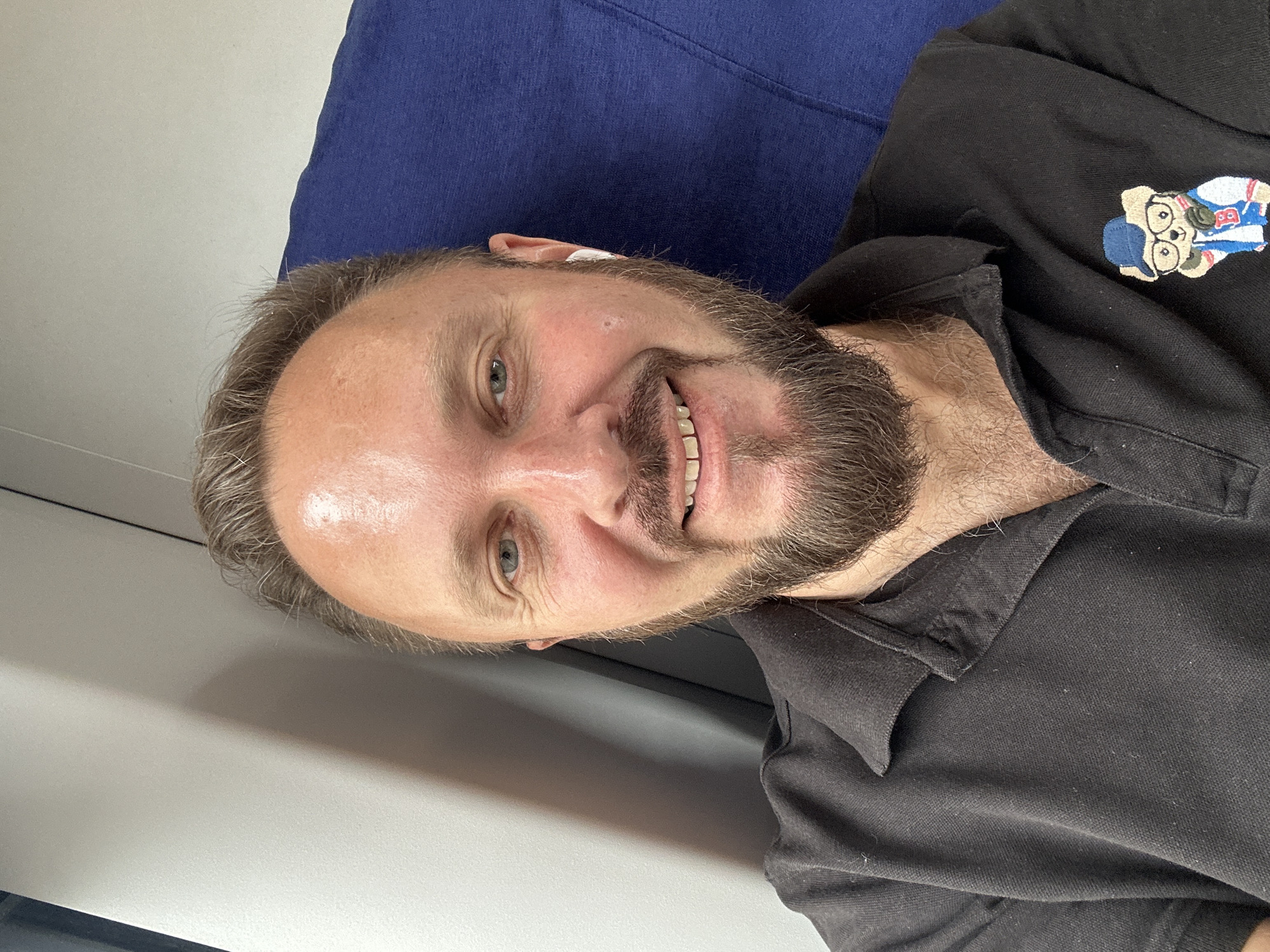 The image is a selfie of a man with fair skin, blonde hair, and a beard, wearing a black polo shirt. He is sitting in what appears to be a train or bus seat, with a blue upholstered seat visible behind him. The man is smiling and looking at the camera. He is wearing white earbuds. On the left side of his polo shirt, there is an embroidered patch of a cartoon teddy bear wearing glasses and a baseball cap. The background suggests an indoor setting with a wall or partition on the left. There is no visible indication of the location being Moscow, Russia.