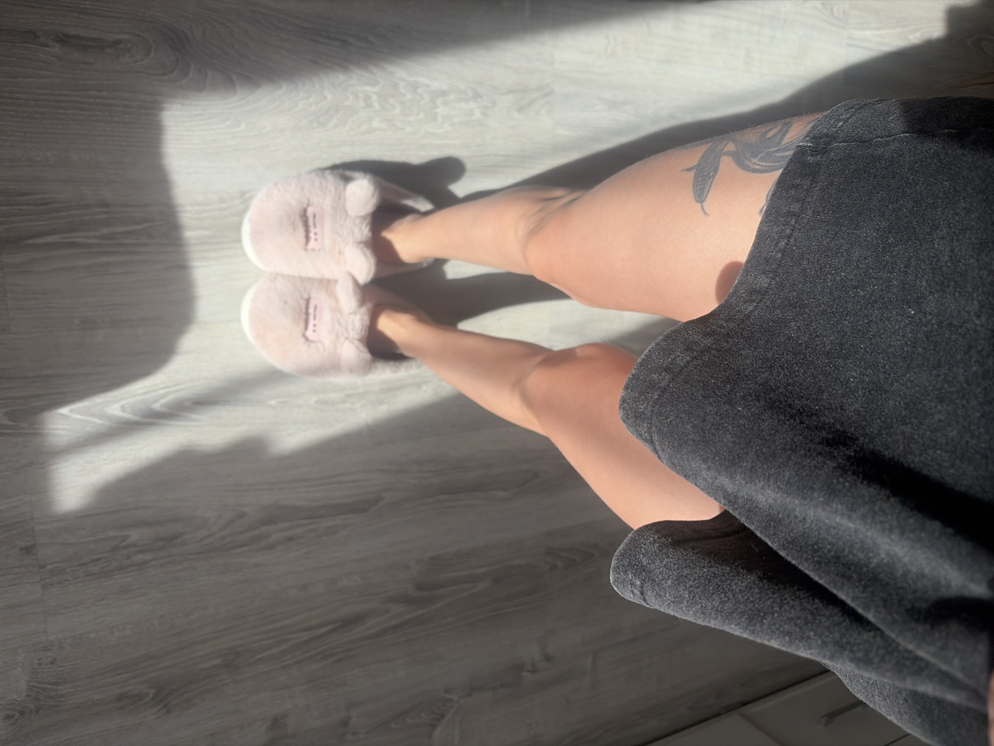A first-person perspective image captures a pair of legs wearing fuzzy, light pink slippers positioned on a grey wood-patterned floor with sunlight casting shadows. The left leg has a tattoo visible on the upper thigh. A dark grey or black garment is partially in the frame on the right side, obscuring part of the view. The setting implies an indoor environment, but there is no evidence to suggest this image was taken in the city of Saint Petersburg, Russia. There are no indicators of a city in the image.