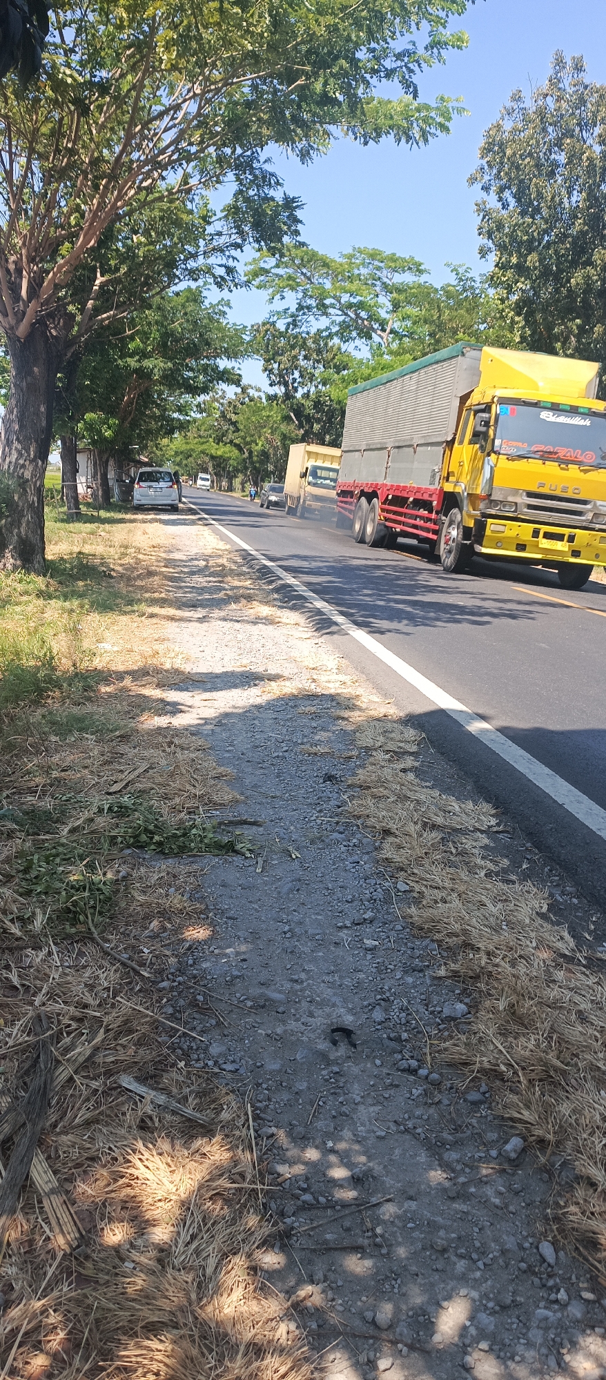 The image captures a sunny day on a road in Landangan, Indonesia, with a distinct focus on the roadside and passing traffic. A yellow truck with a grey cargo box is prominently featured on the right side of the road, moving forward. To the left of the truck, a lighter-colored truck is also in motion, emitting dark exhaust fumes. Further down the road, several other vehicles, including a white car and a bus, are visible. The roadside on the left is characterized by dry grass and a gravel path, with a large tree providing shade in the foreground. A small white structure, possibly a building or a roadside stall, is situated behind the parked white car. The environment appears dry and natural, with trees lining the road and a clear blue sky overhead.