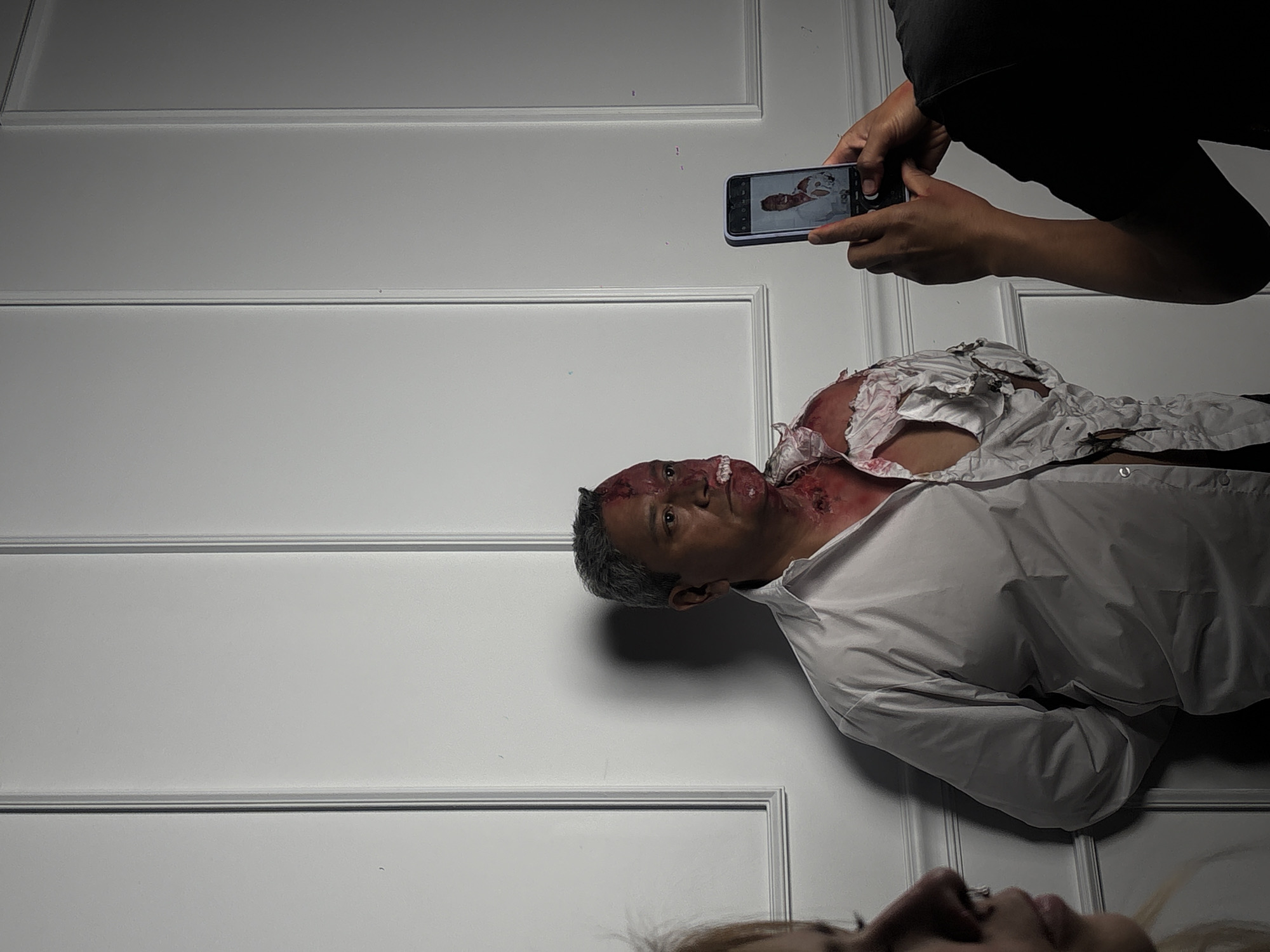 The image depicts an apparent crime scene. A man with apparent head trauma and blood staining his white shirt is lying next to a white wall. Another person is taking a photo of the man with a mobile phone. The context suggests a forensic or investigative setting. The precise location in Mexico City, Mexico, is not determinable from the provided image.
