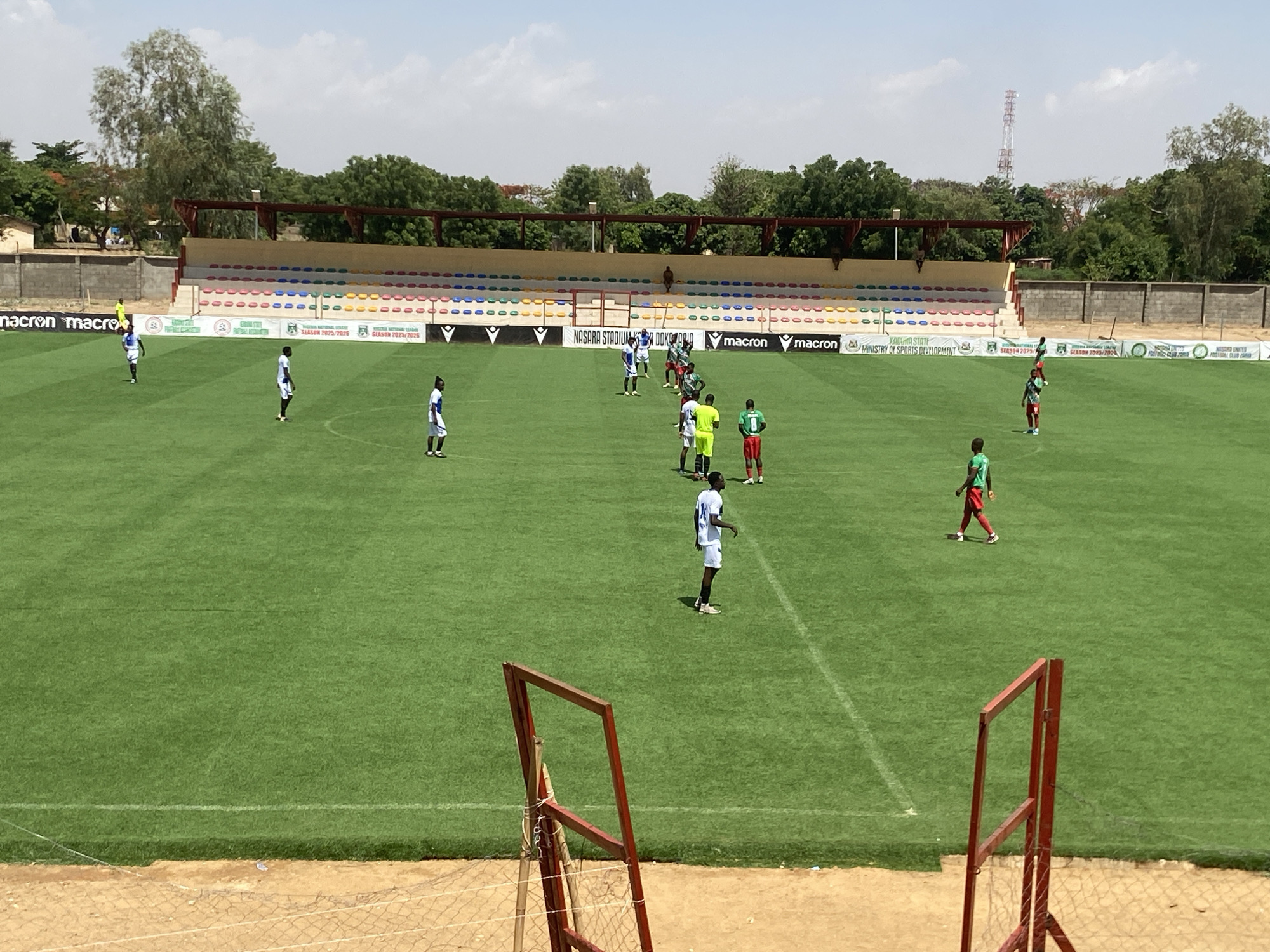 The image depicts a daytime scene at Nasarawa Stadium in Tudun Wada, Nigeria. A soccer match is underway on a well-maintained green field. Players in blue and white uniforms are positioned across the field, along with players in green and red uniforms. A goalkeeper is visible near the center of the field. The stadium seating behind the field is mostly empty. Banners bearing the "macron" logo and the logos of the Kaduna State Ministry of Sports Development are visible. A radio tower is visible in the background. A red frame of the goalpost is in the foreground.