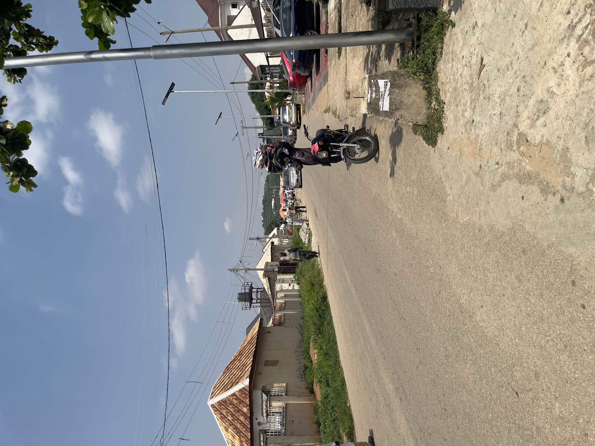 The image depicts a street scene in Abuja, Nigeria, under a clear blue sky with scattered clouds. A paved road occupies the foreground and extends into the distance. On the left side of the road, a row of single-story buildings with rusted, corrugated iron roofs is visible, bordered by overgrown grass. Several utility poles with numerous power lines are strung across the scene.
On the right side of the road, a concrete sidewalk is present, with a light pole and some overgrown vegetation. A motorcycle is parked on the sidewalk, and a person is standing next to it, carrying a large stack of folded items on their head. Further down the street, several cars are parked along the side of the road, including a red, a dark blue, and a silver sedan. More buildings, some with modern architectural styles and tiled roofs, are visible in the background, along with a water tower and distant hills. People are walking on the sidewalk and the road in the middle ground. The overall impression is of a residential or mixed-use urban area in a somewhat dry climate.