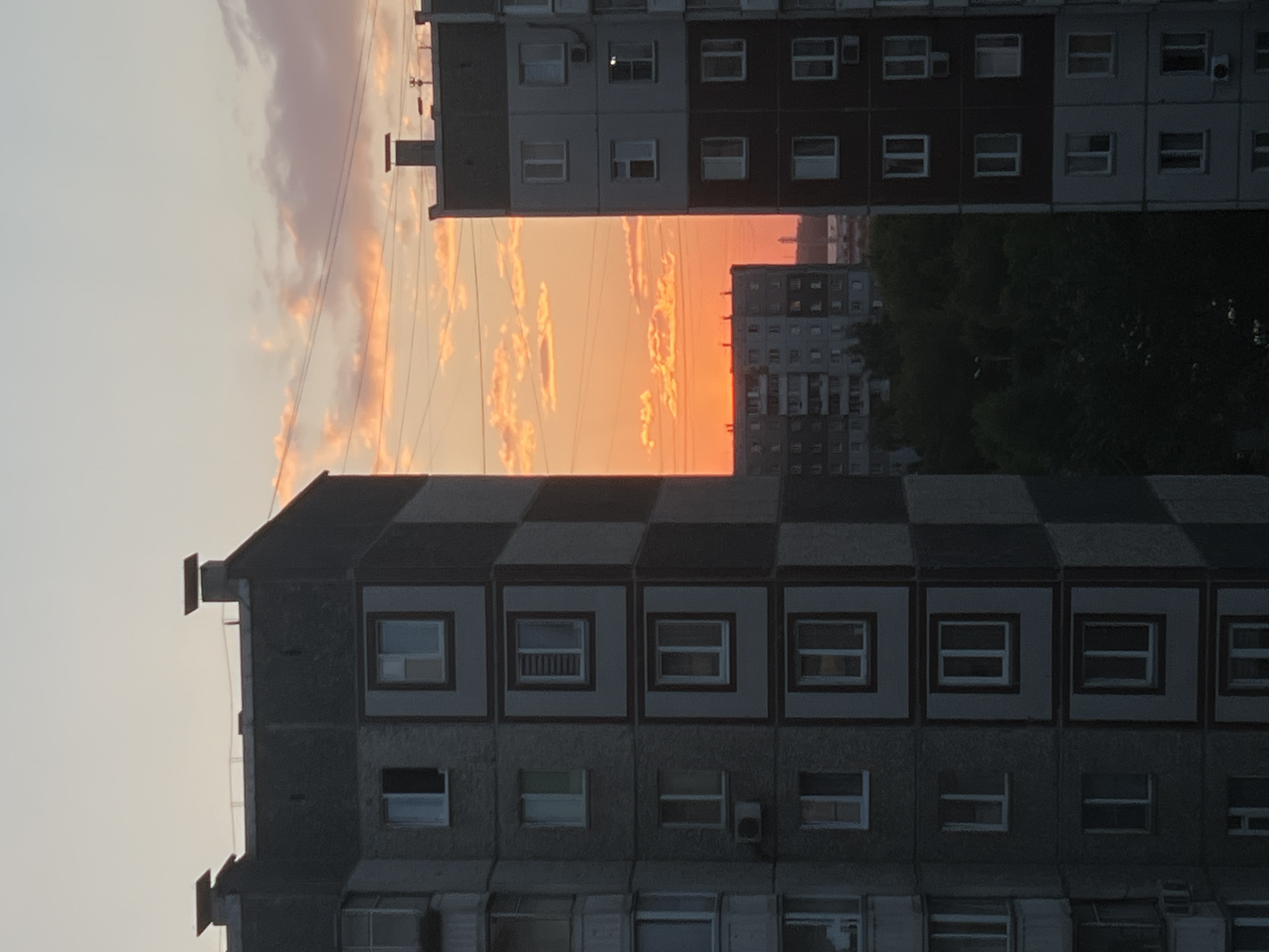 The media file depicts a view of residential buildings in Krasnoyarsk, Russia, during sunset. The sky is filled with warm orange and pink hues, indicating the sun is low on the horizon. Several multi-story apartment buildings are visible, some with dark facades and others with lighter, checkered patterns. Windows are scattered across the buildings, with some appearing to be lit from within. Power lines are visible in the sky, stretching between structures. The overall scene suggests an urban environment during the evening. There is no visible text in the image.