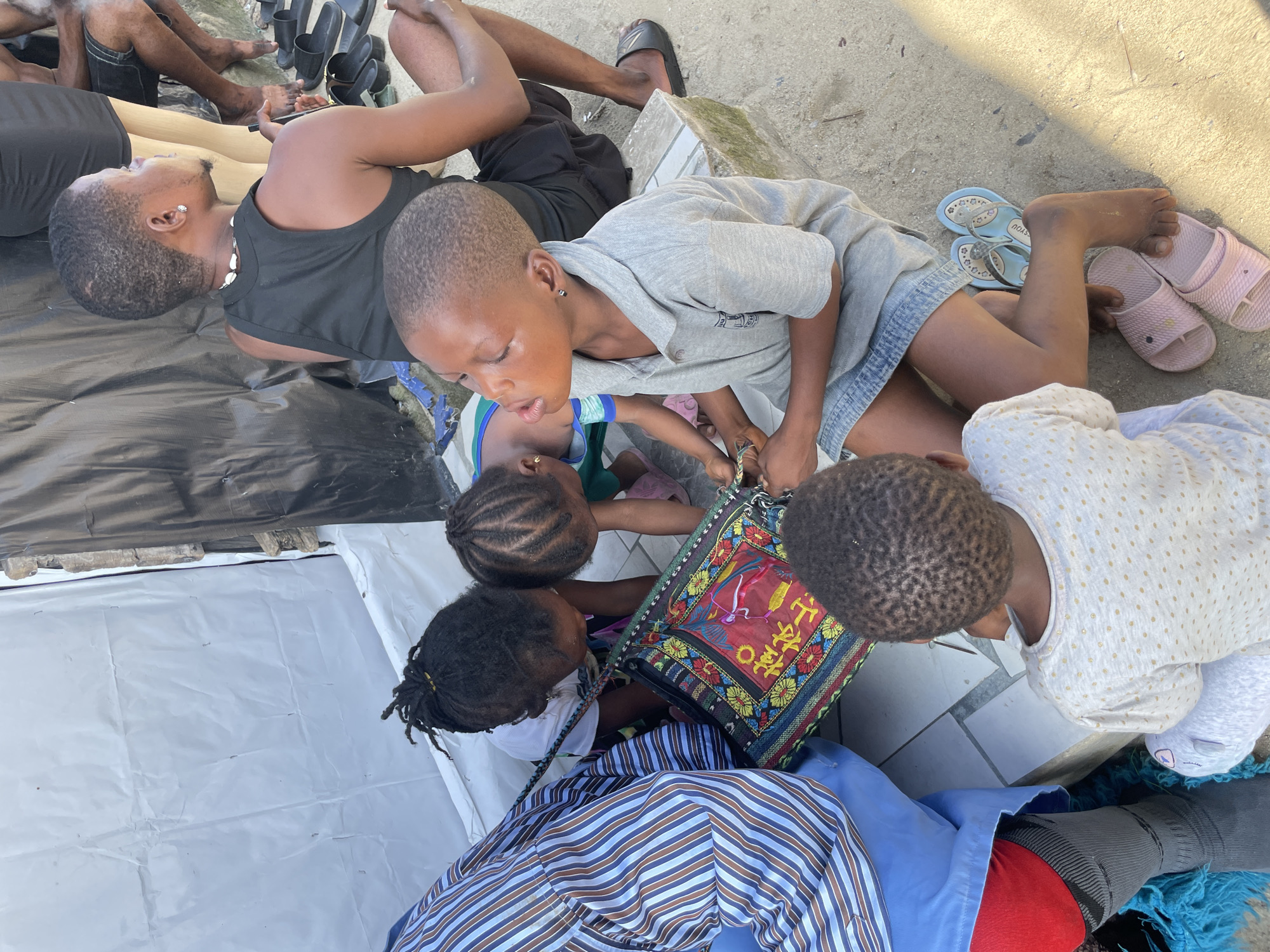 In Iranla, Nigeria, a group of people, including children and adults, are gathered outdoors. One adult, wearing a black tank top and necklaces, sits with their back to the camera, looking to the right. Another adult's legs are visible on the right, wearing black shorts and flip-flops. In the foreground, several children are interacting. One child, wearing a grey collared shirt and denim shorts, is holding a patterned bag with red and yellow accents. Another child, wearing a white shirt with gold polka dots, is seated facing away from the camera, looking down. Two other children are leaning over the patterned bag, their heads close together. The environment appears to be sandy with some tiled surfaces and a white and black tarp-like structure in the background. Various footwear, including flip-flops and sandals, are scattered around.