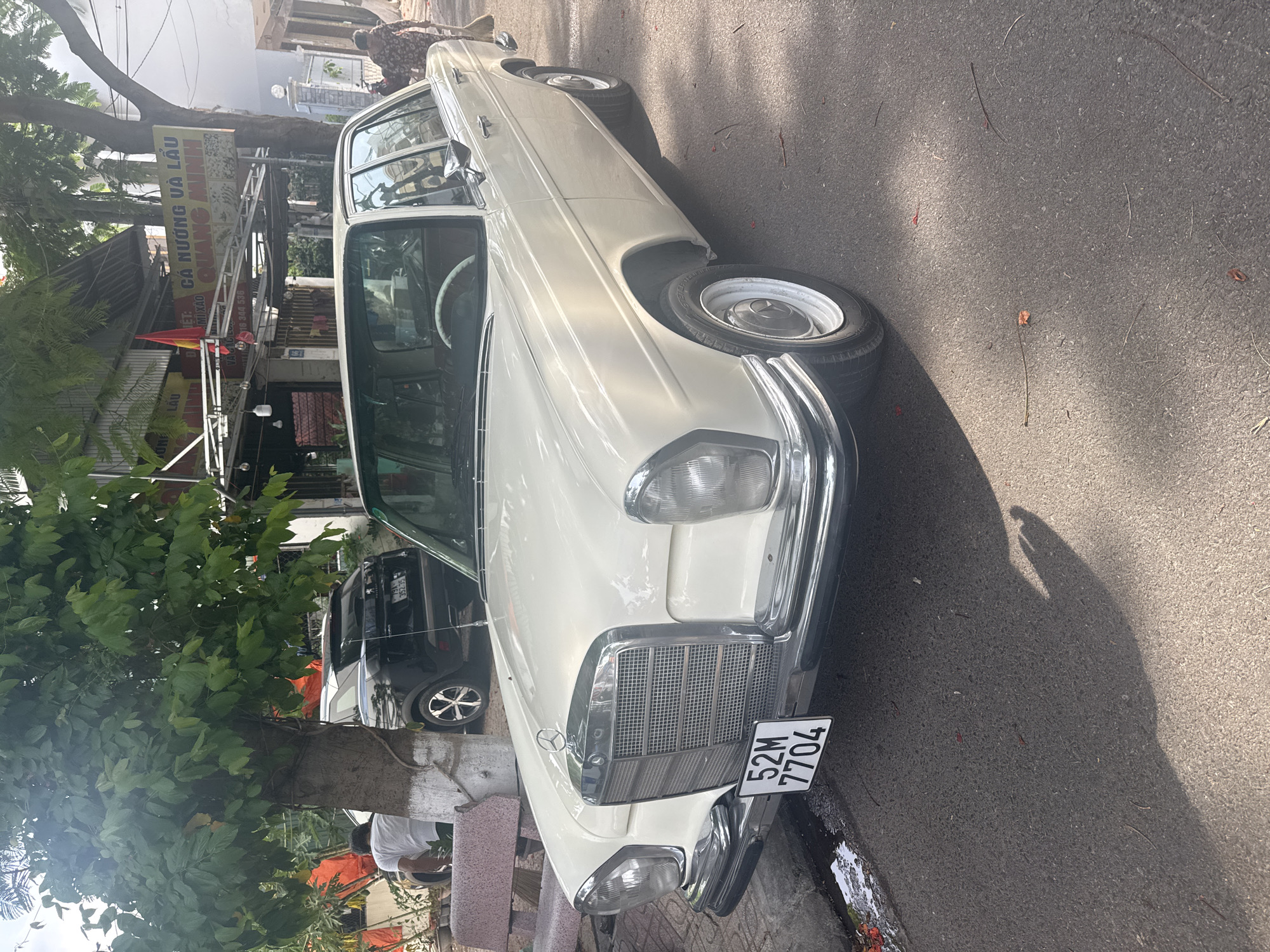 A light-colored, vintage Mercedes-Benz sedan is parked on a paved street in Bac Nha Trang Ward, Vietnam. The car is facing left, with its front end slightly angled towards the viewer. A license plate that reads "52M 7704" is affixed to the front bumper. To the left of the car, a black SUV is visible, partially obscured by a tree with green foliage. In the background, a building with a yellow sign displaying Vietnamese text is situated under an awning. A person in a patterned dress is walking away from the camera to the right of the Mercedes. The street is paved and appears to be in a somewhat urban environment, with shadows cast across the ground by the surrounding trees and structures.