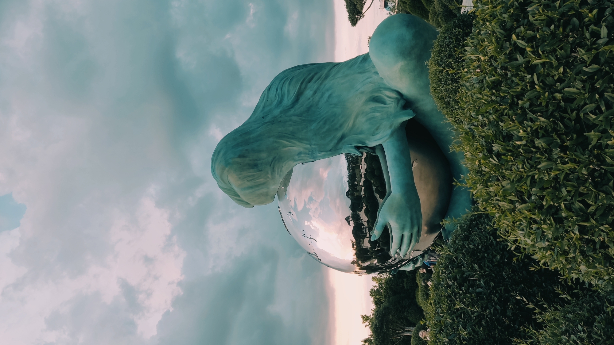 The image depicts a large, teal-colored statue of a mythical creature, possibly a mermaid or siren, embracing a giant, reflective sphere. The statue's hair flows out behind it, and its hands cradle the sphere. The sphere reflects the surrounding environment, showing trees, bushes, and a cloudy sky with hints of pink and orange from what appears to be a sunset or sunrise. The statue is situated in a landscaped garden with manicured green bushes in the foreground. In the background, there are more trees and some distant structures. The sky is filled with dramatic, dark clouds tinged with lighter hues. The overall atmosphere is serene and slightly melancholic. This statue is located in Krasnodar, Russia, and is known as "The Mermaid" or "Mermaid of the Kuban."