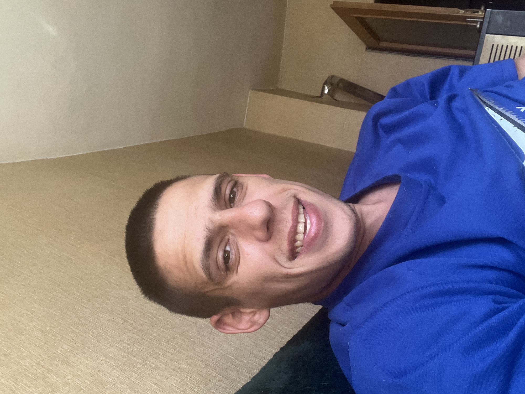 A man with short, dark hair and a slight tan is smiling towards the camera. He is wearing a bright blue t-shirt. A pair of scissors is visible near his shoulder. The background includes a beige wall and what appears to be a shelf unit. The location is not discernible from the image provided, and I cannot confirm that it is in Sofia, Bulgaria.