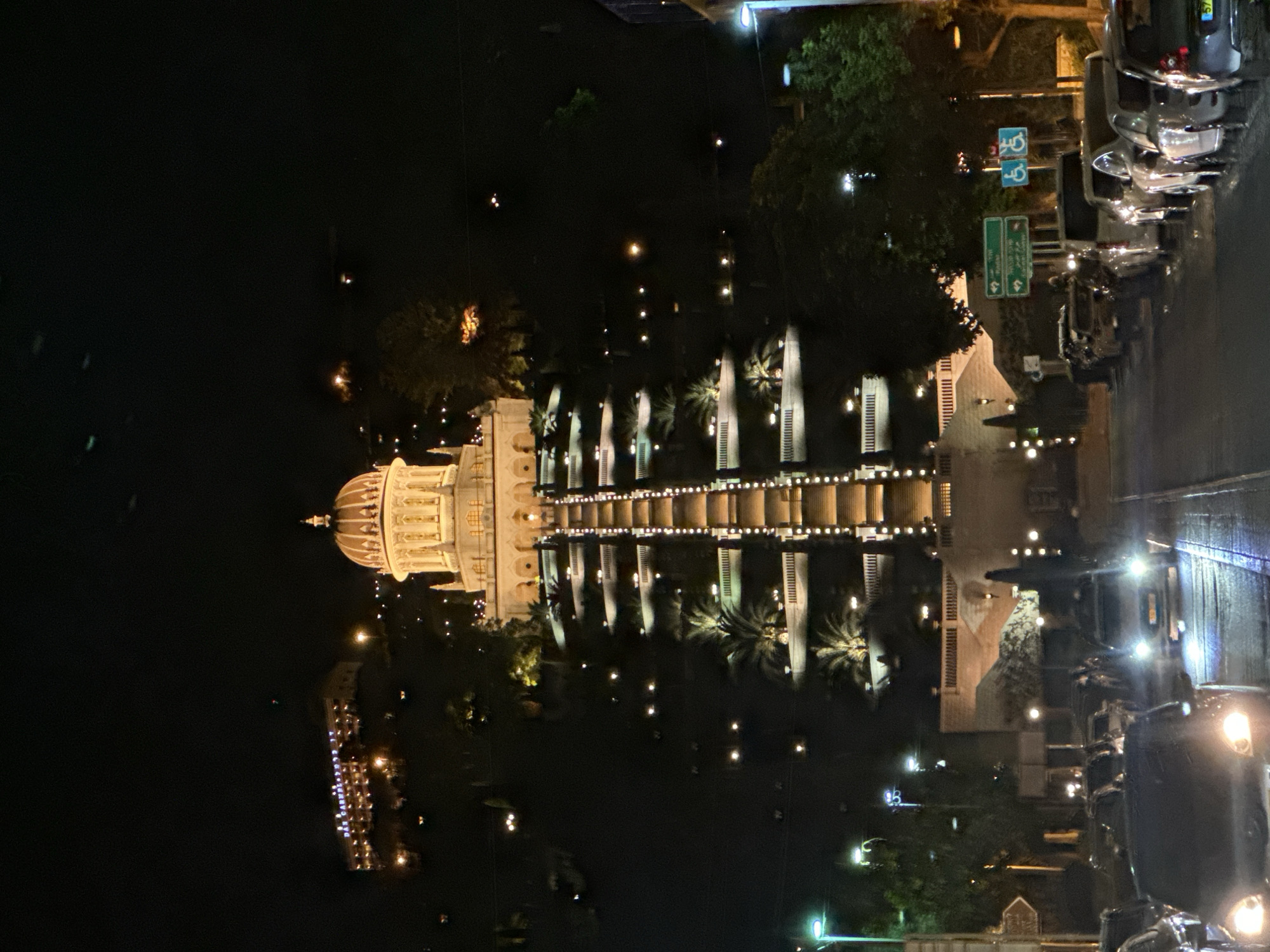 This media file captures a nighttime scene of the Baháʼí World Centre in Haifa, Israel. The primary subject is the illuminated dome and arcades of the Terraces. The building, bathed in warm light, is reflected in the dark water, creating a symmetrical and serene ambiance. Palm trees are visible, silhouetted against the lights and the night sky. Several cars are parked along a street to the right of the image, their headlights and taillights reflecting on the wet pavement, suggesting recent rain or mist. The sky is dark, indicating nighttime, and the overall mood is peaceful and contemplative. Two green road signs with white text are visible near the cars, one showing directional arrows. Above them, two blue signs with international symbols for accessibility are present. The image's orientation is rotated, with the dome appearing at the bottom left and the street scene at the top right.