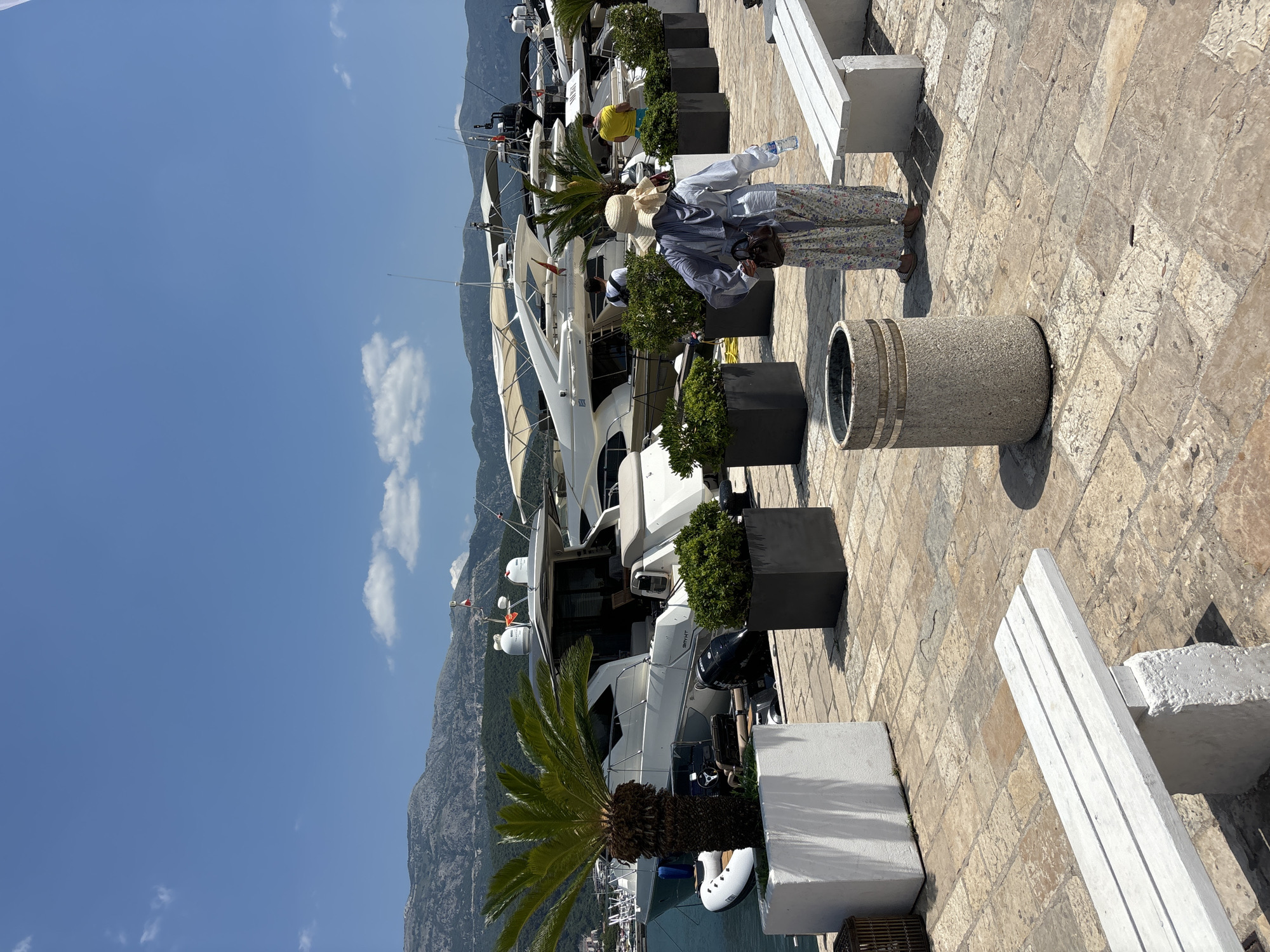 A sunny day in Budva, Montenegro, with a view of a marina and moored yachts. The foreground features a paved promenade with several white benches and planters. One planter contains a palm tree, while others have dense green bushes. A concrete trash receptacle is also visible. Two people are on the promenade; one, seen from behind, is wearing a wide-brimmed hat, a long-sleeved shirt, and patterned pants, and is holding a bottle of water. Another person, wearing a yellow shirt, is standing near the yachts in the midground. The midground is dominated by several white yachts, some with canopies, docked in the marina. In the background, rolling hills covered in vegetation rise against a clear blue sky with scattered white clouds.