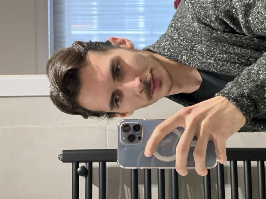 An individual is depicted taking a mirror selfie in an indoor setting. The subject is a male with dark brown hair, styled with volume, and a dark brown mustache. He wears a dark, possibly black, round-neck t-shirt under a grey, textured, flecked cardigan or jacket. His right hand holds a light blue/grey iPhone, featuring a clear case and a visible white MagSafe ring, indicating the method of image capture. In the background, to the left, a black, modern heated towel rail with horizontal bars is visible against a light-colored wall. To the right, a reflection of a window with vertical blinds or curtains is present. The specific location within the city of Tbilisi, Georgia, cannot be determined from the visual evidence provided.