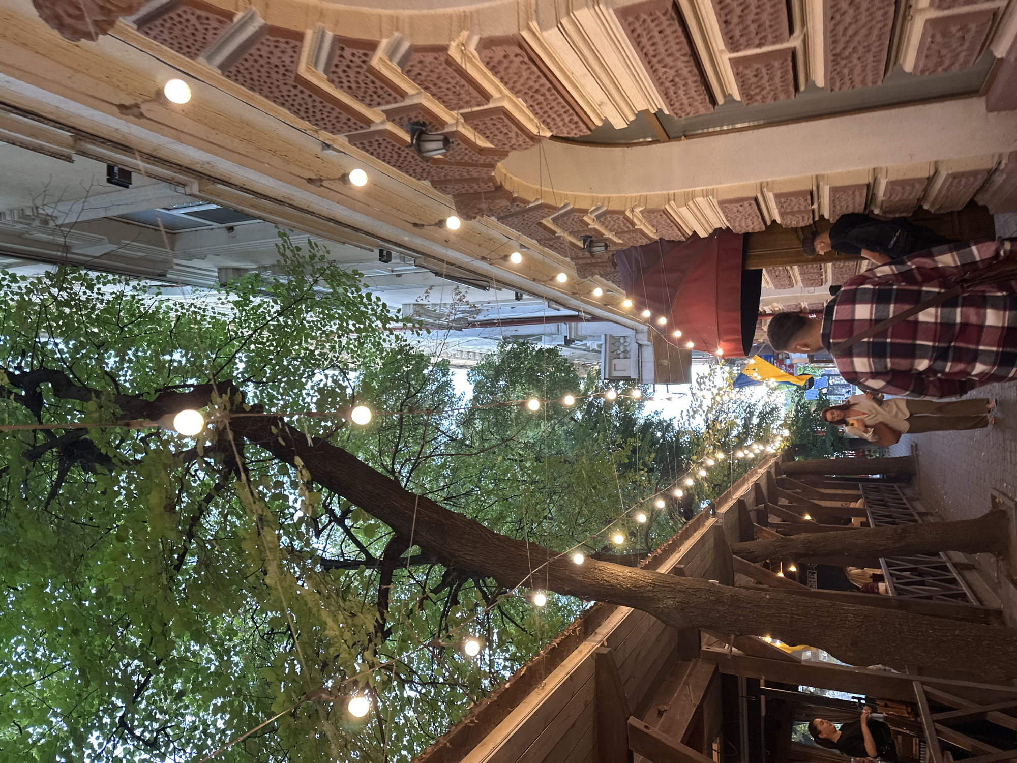 The image depicts an outdoor seating area of a restaurant or cafe on a cobblestone street lined with trees and string lights. The building on the right side of the street has an ornate facade with brickwork and decorative elements. People are visible, including a woman in a light-colored blazer and trousers walking down the street, and a person in a plaid shirt walking away from the camera. On the left, a covered outdoor seating area is visible with people seated at tables. The trees are leafy and green, suggesting a warm season. String lights are strung between the trees and along the building, creating a festive atmosphere. The overall scene suggests an evening setting, possibly twilight. The location is identified as Ternopil, Ukraine.