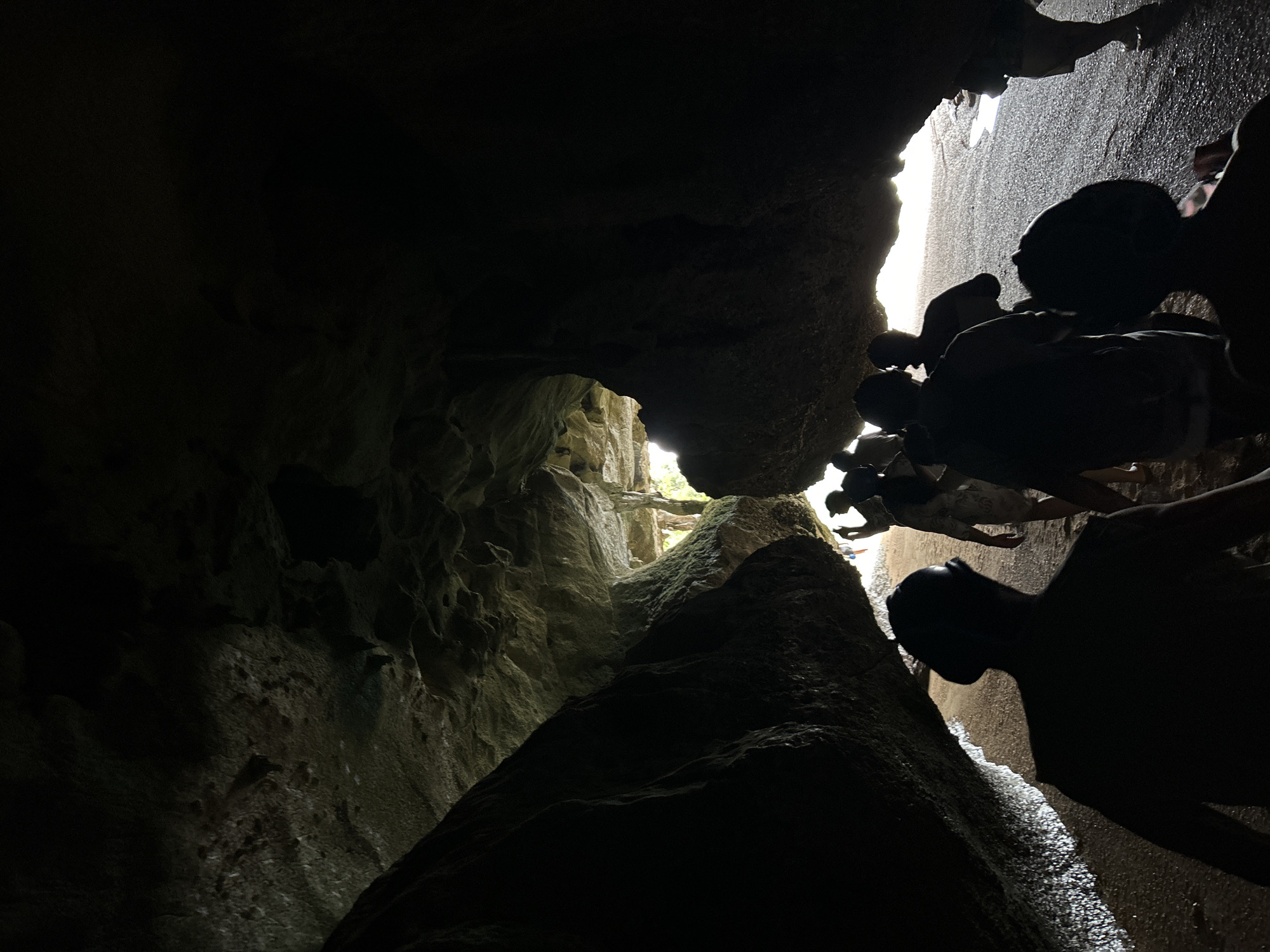 The image depicts a group of people walking through a narrow, dimly lit cave opening, presumably on a beach in Krasom, Thailand. The cave walls are rough and textured, with some appearing to be made of rock and others possibly of eroded sand or soil. Sunlight streams in from the opening ahead, illuminating the sandy ground and casting shadows. The people are silhouetted against the light, making it difficult to discern individual features. They appear to be walking in a single file line, suggesting they are on a guided tour or exploring together. The overall atmosphere is one of mystery and adventure, with the natural beauty of the cave being the main focus.