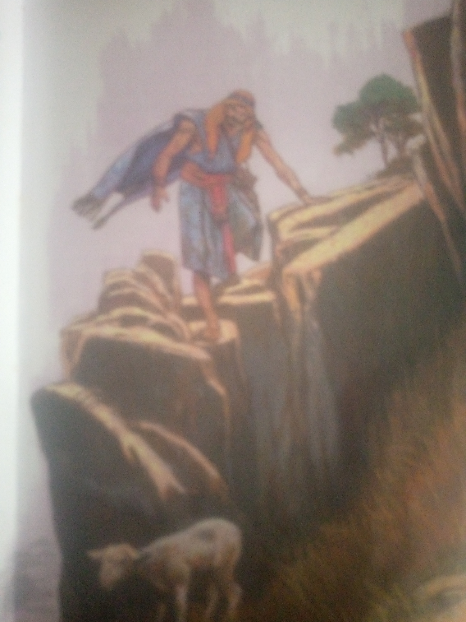 A watercolor painting depicts a figure, likely a man, standing on a rocky cliff. He wears a blue robe draped over one shoulder and a red sash around his waist. His hair is blonde, and he has a beard. His body is angled as if he is leaning forward or stepping. Below him, nestled amongst the rocks, is a small, light-colored lamb. The scene is set in a rugged, natural landscape with large rock formations and a distant tree. The overall impression is one of a dramatic or spiritual moment, possibly evoking themes of protection or sacrifice, given the presence of the lamb. The lighting suggests it could be daytime, but the muted colors and hazy atmosphere might imply dawn or dusk. No text is visible in the image.