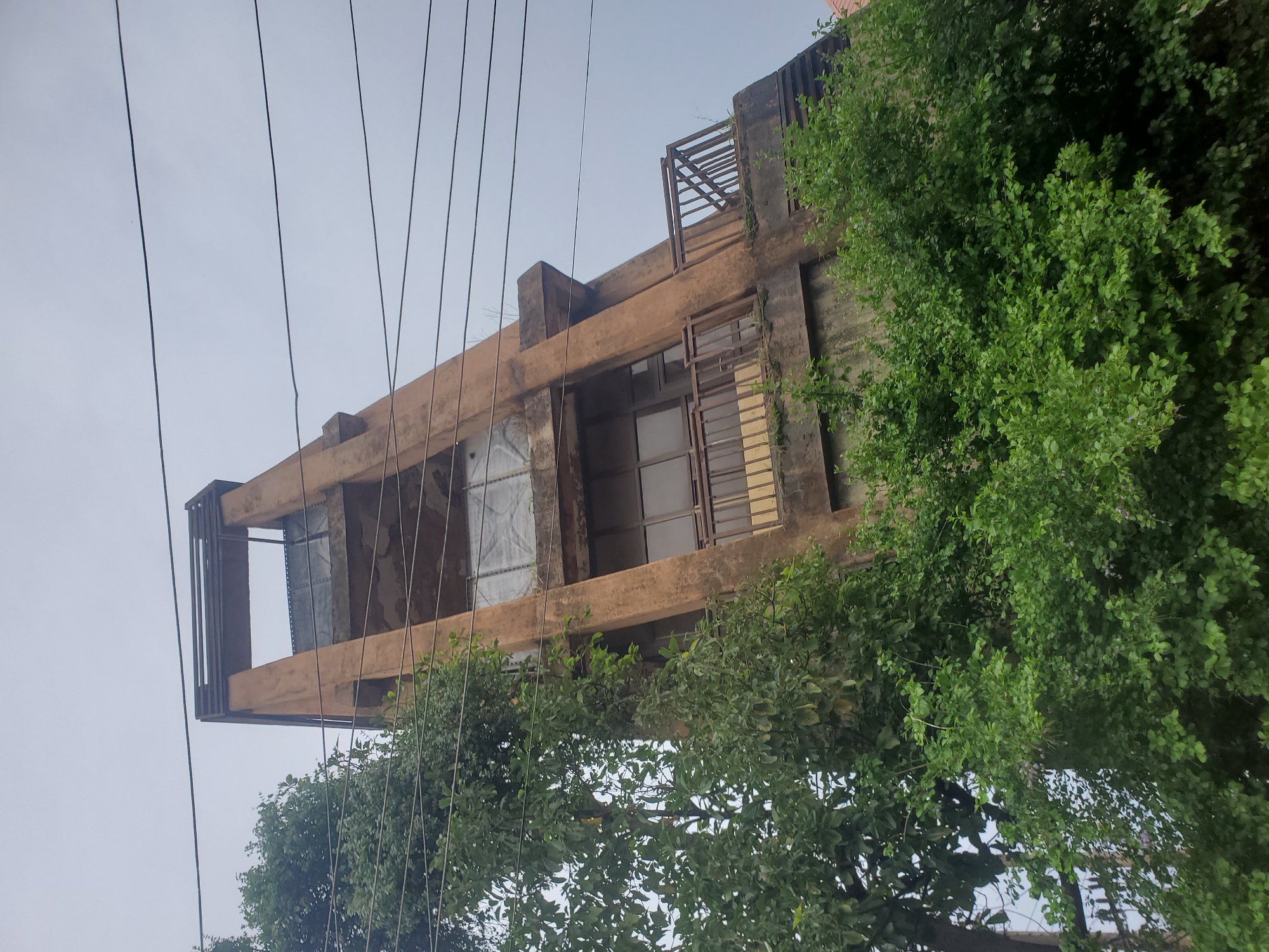 A tilted, multi-story concrete structure, possibly an abandoned water tower or building, stands against an overcast sky in Jos, Nigeria. The structure is weathered and appears to be stained with age and possibly water damage. It is flanked by lush green foliage, with trees and bushes partially obscuring the lower levels. Electrical wires are visible crisscrossing the sky above the structure. The building features several levels with windows, some of which are covered with translucent sheeting, and a balcony with a metal railing on one of the upper floors. The overall impression is one of decay and overgrowth, suggesting a long period of disuse.