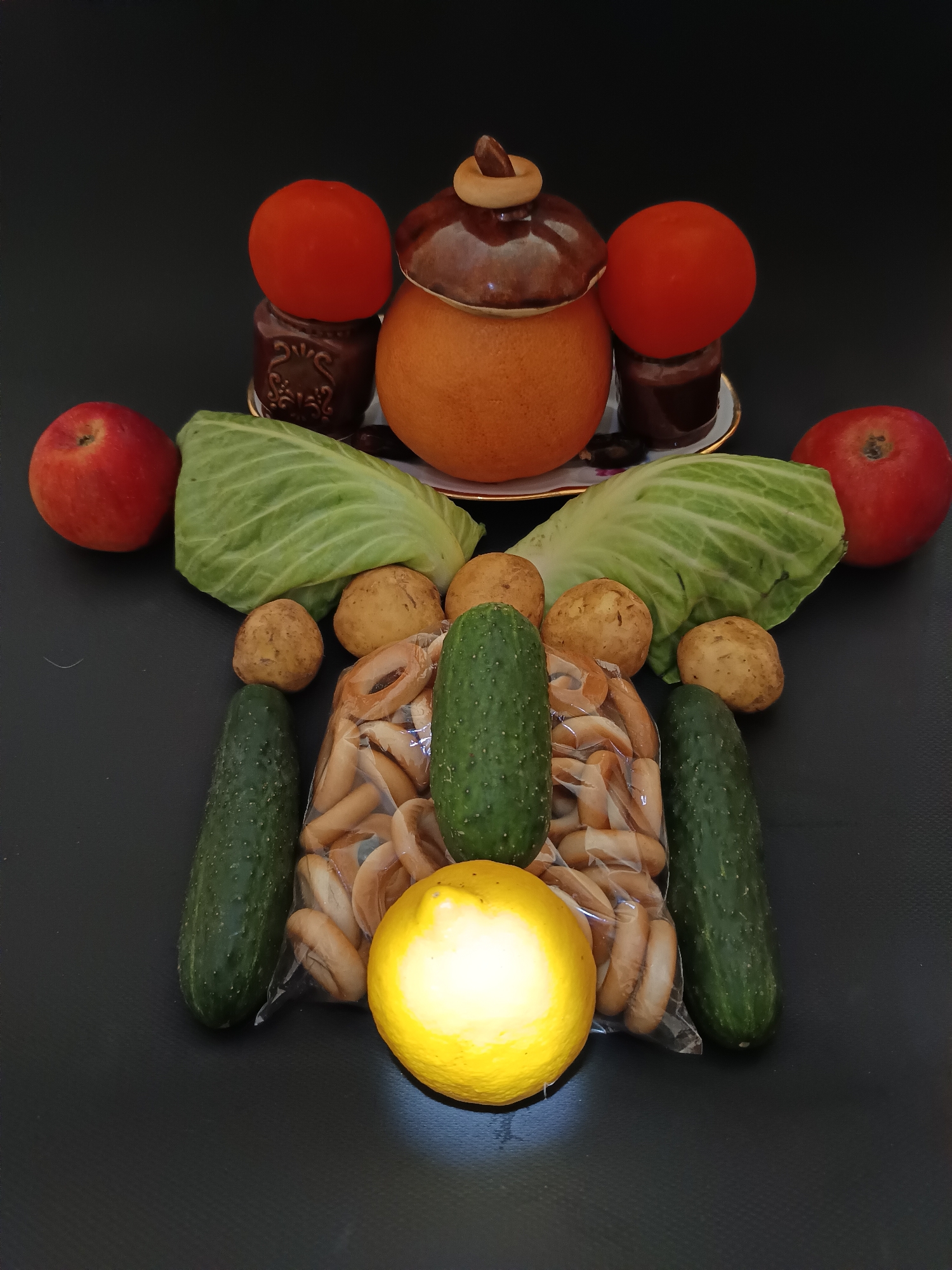 A still life arrangement of food items is displayed against a dark background. The subjects include two red apples, two heads of cabbage, several potatoes, two cucumbers, a bag of bread rings, and a bright yellow lemon. At the top of the arrangement, there is a ceramic pot with a lid, flanked by two tomatoes and two small jars of dark liquid. The scene is lit from the front, casting shadows and highlighting the textures of the produce. The overall impression is one of abundance and natural bounty. The setting appears to be indoors, with the dark background suggesting a studio or controlled environment. No text is visible in the image.