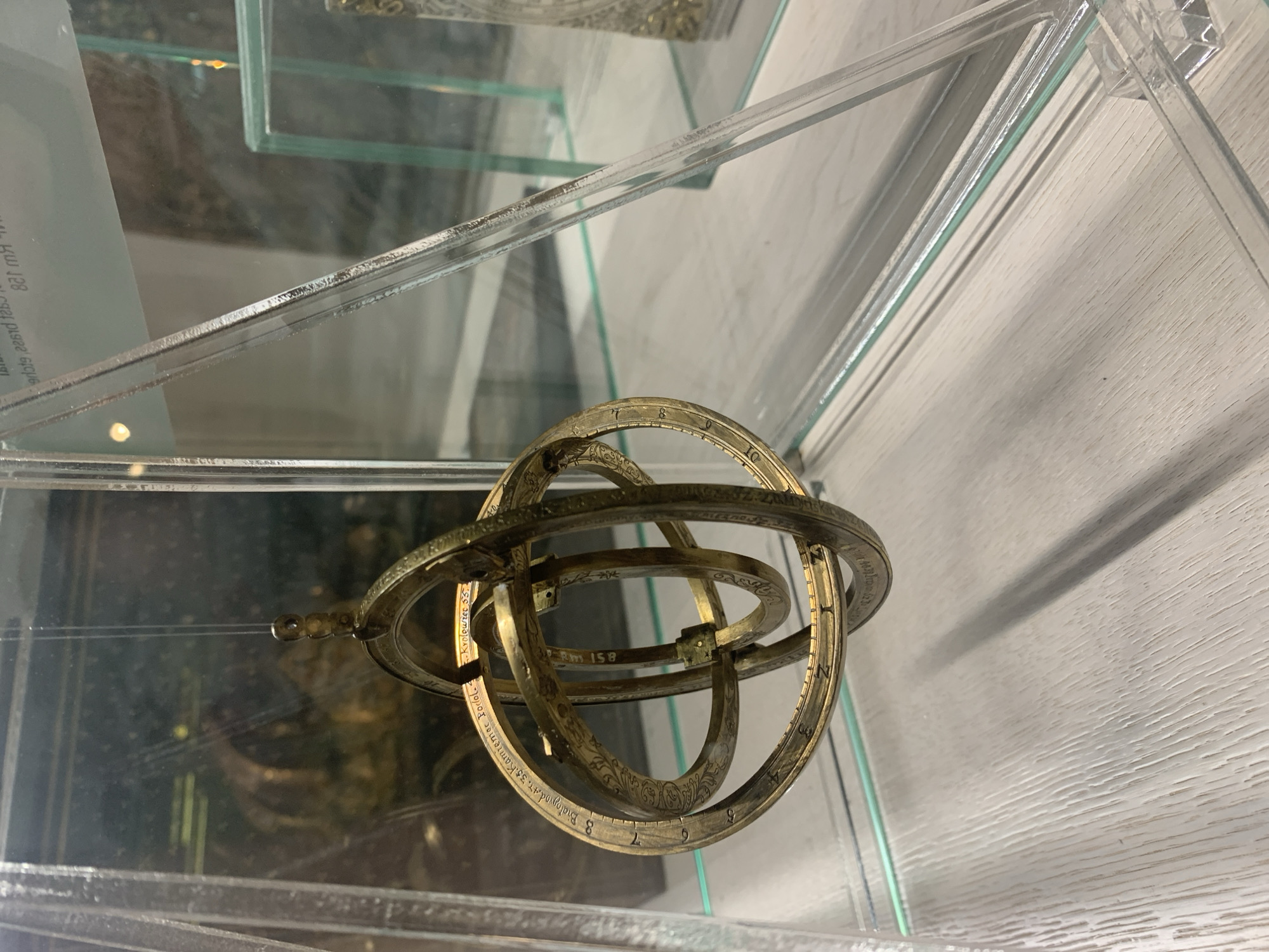 An armillary sphere, a spherical astronomical instrument, is displayed in a glass case. The sphere, made of brass, features multiple concentric rings with engraved markings and patterns. It is suspended by a thin wire from the top of the case. The background shows reflections of other museum exhibits and hints of text. The case itself is made of clear acrylic and stands on a light-colored wooden surface. The armillary sphere is the primary subject, centrally positioned and in focus. The overall scene suggests a museum or exhibition setting. This object is located in the National Museum in Poznań, Poland.