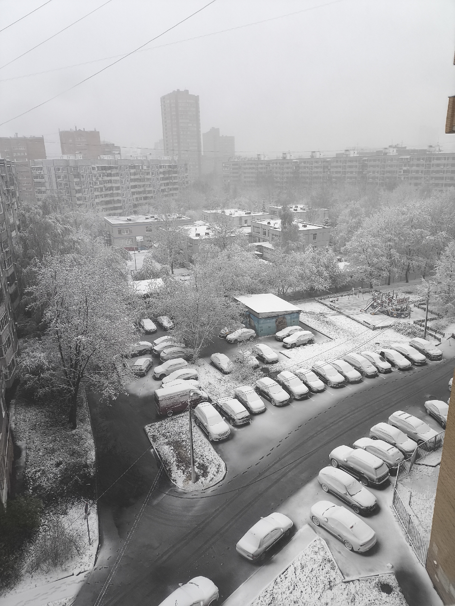 The image depicts a snow-covered urban landscape, likely a residential area in Khimki, Russia, characterized by buildings with dense housing blocks, trees, parked vehicles, roads, and a children’s playground, all covered in a blanket of snow. The parking lot is occupied by dozens of cars. The image appears to be taken from an elevated position, offering an overview of the scene. The heavy snowfall obscures the visibility of distant objects, creating a hazy atmosphere.