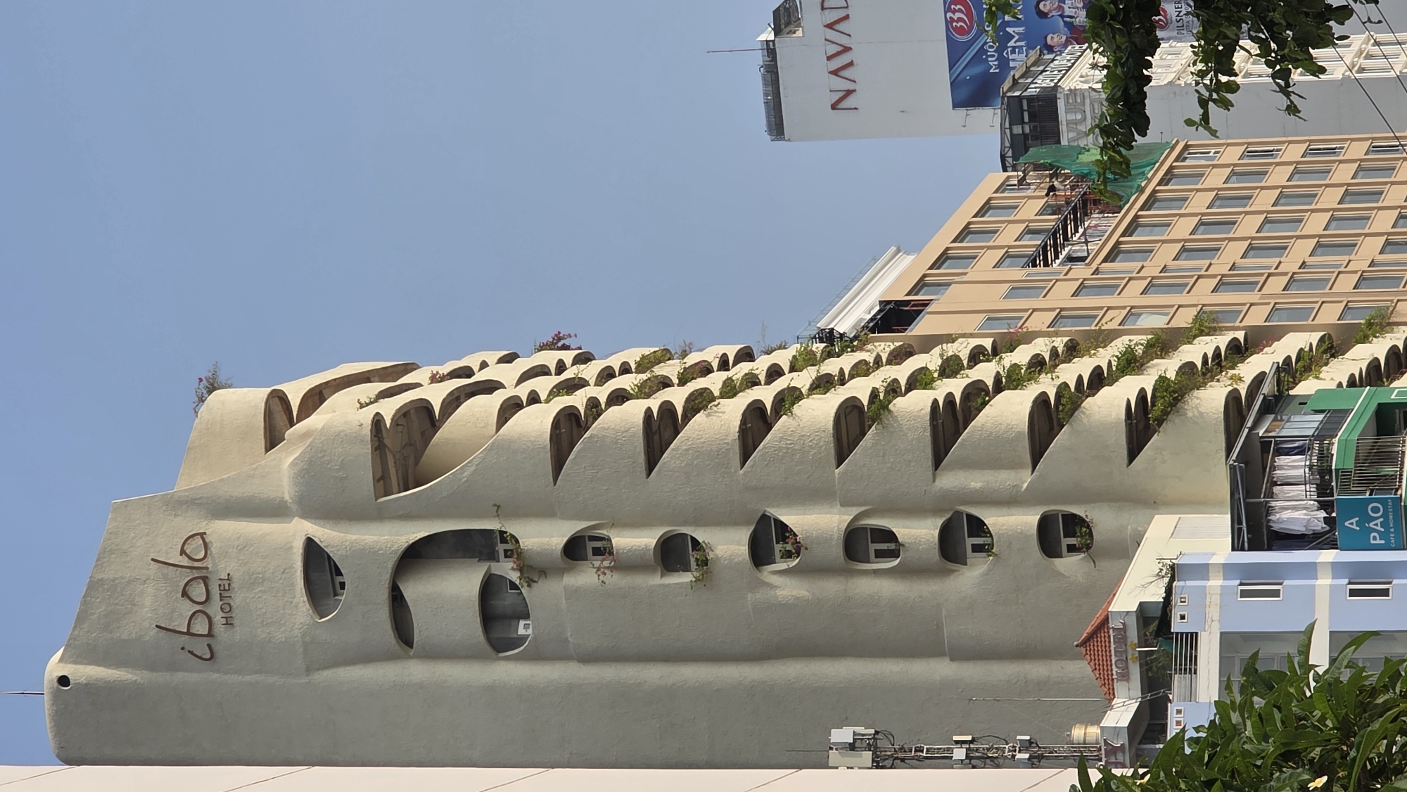 The image depicts a close-up view of the 'ibala hotel' located in Bắc Nha Trang, Vietnam. The hotel's architecture is characterized by a distinctive, wavy facade with rounded cutouts forming arch-like structures on its upper levels. Small plants are visible atop and around these arches. Lower sections of the facade feature rounded window openings. A sign displaying 'ibala HOTEL' is seen. Behind the hotel, taller buildings are visible. One building has a light brown facade with rows of windows and another building has a large billboard. The sky is clear and blue. The shot is angled upwards.