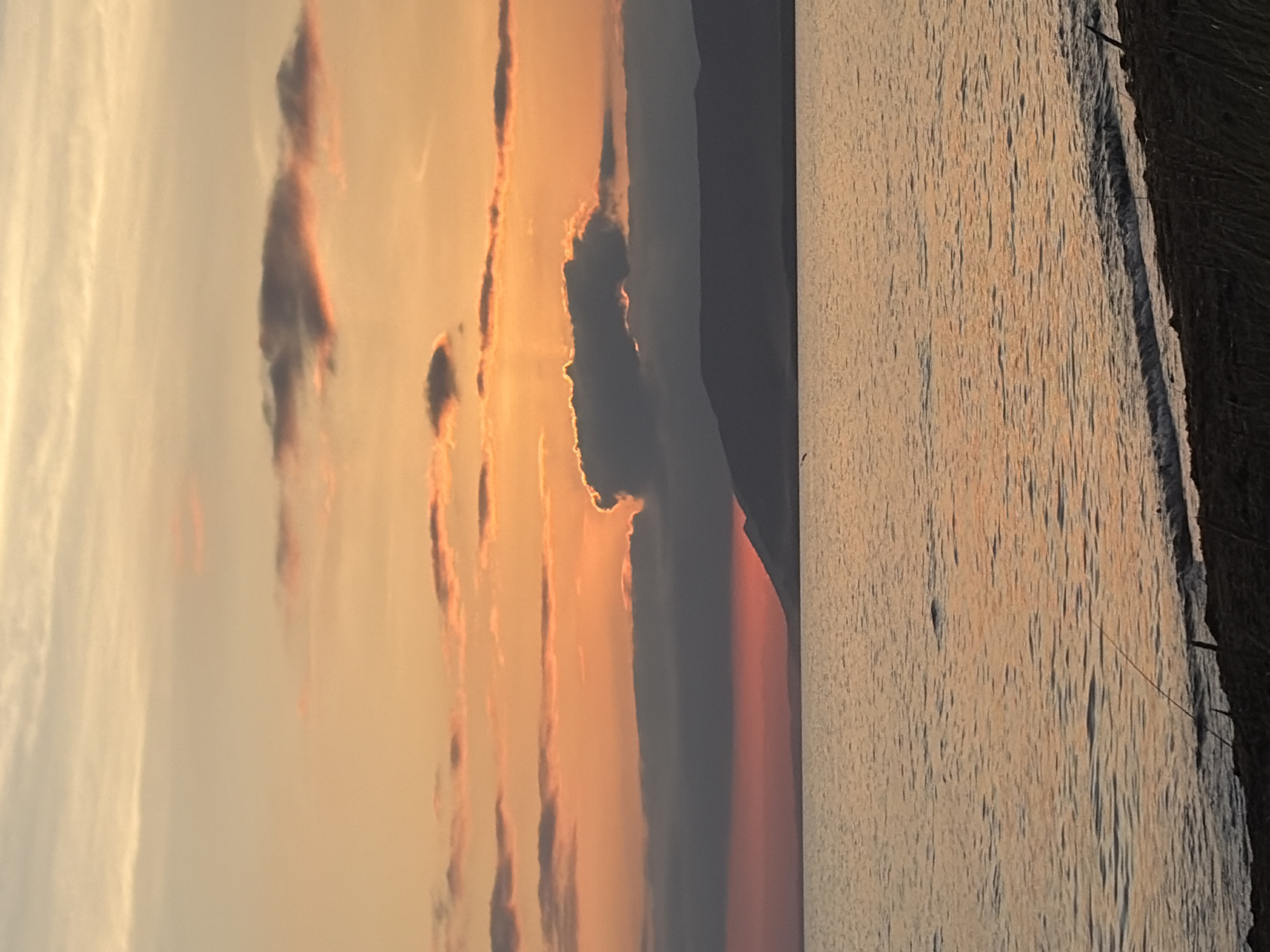 The image depicts a serene sunset over the body of water in Ozerskoe, Russia. The sky is ablaze with hues of orange and pink, with wispy clouds scattered across the horizon. The sun, partially obscured by a dark cloud formation, casts a warm glow on the water's surface, creating ripples and reflections. A silhouetted mountain range forms the backdrop, adding depth and grandeur to the scene. The foreground features a sandy shore with sparse vegetation, hinting at a natural, undeveloped environment. The overall atmosphere is tranquil and picturesque, evoking a sense of peace and natural beauty.