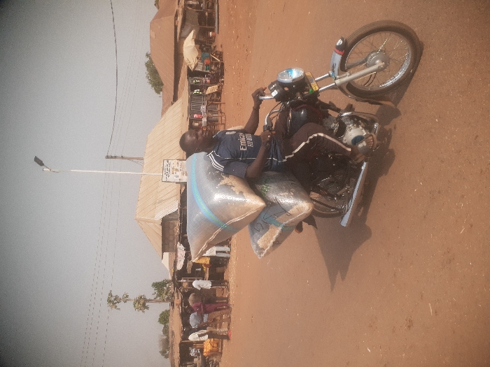 The image displays an outdoor street scene in Gwadanye, Nigeria, under daylight conditions. In the foreground, a dark-skinned male, wearing a navy blue short-sleeved t-shirt with "Emirates Fly Better" and dark shorts with white stripes, is seated on a black motorcycle. The motorcycle's rear is heavily loaded with two large, light-colored sacks, appearing to contain granular material, secured one atop the other. The rider's hands are positioned on the handlebars, and his head is turned slightly to his left (viewer's right). The ground is an unpaved, reddish-brown dirt surface, with shadows cast by the motorcycle and rider.
In the mid-ground, to the viewer's left, a group of approximately 5-7 dark-skinned individuals is visible, congregating near simple structures featuring corrugated iron roofs, indicative of market stalls or shops. To the viewer's right, more similar structures with corrugated iron roofs and a white market parasol are present. A tall, slender white street light pole, equipped with a modern LED luminaire, stands centrally in the background. Overhead utility wires are discernible, and several sparse trees with light green foliage punctuate the background against a pale, possibly hazy sky.