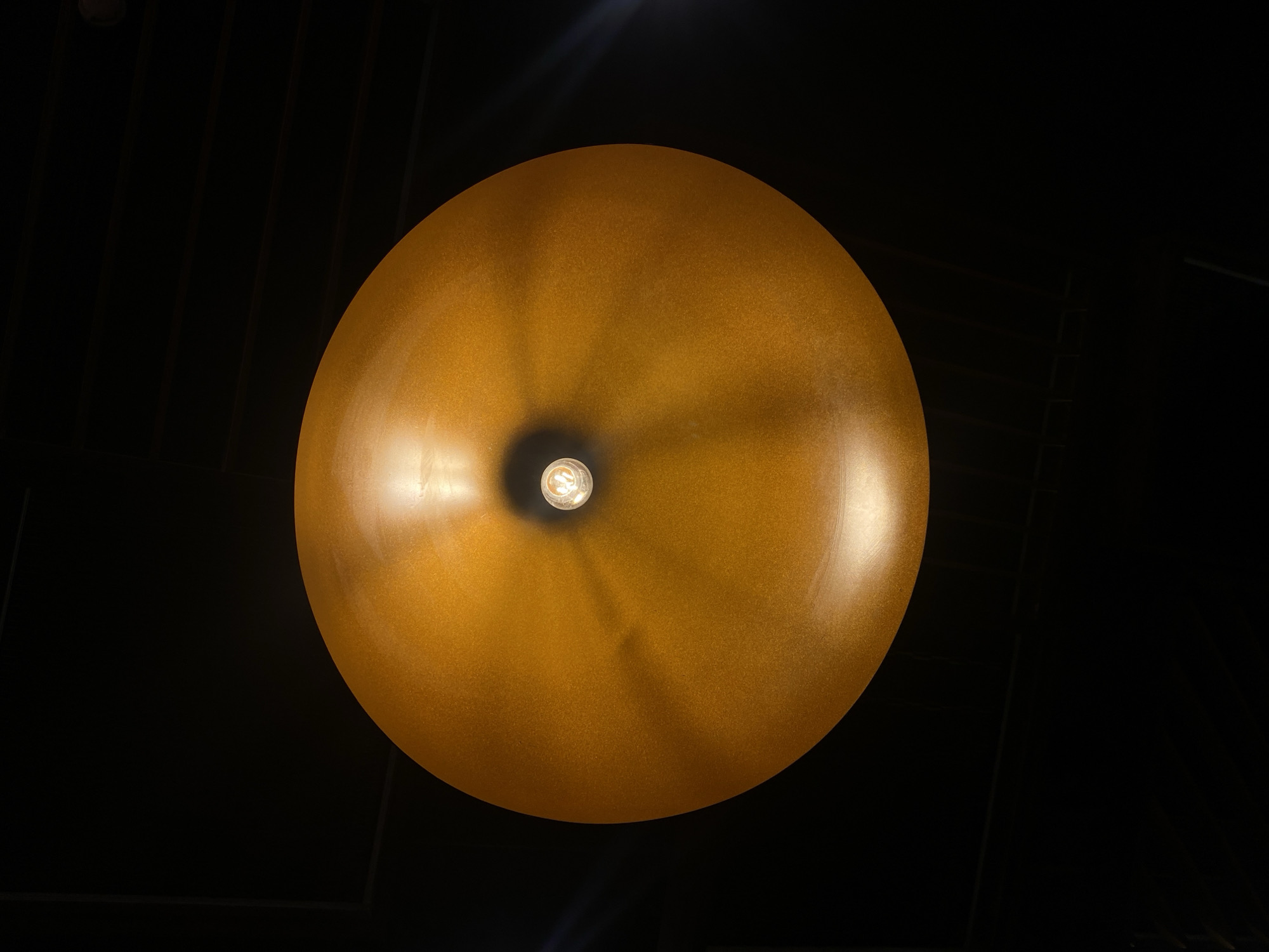 The image shows a golden, spherical lampshade illuminated from within by a bulb. The background is a dark ceiling, possibly with linear elements. There's no information to deduce that the location is Kazan, Russia.