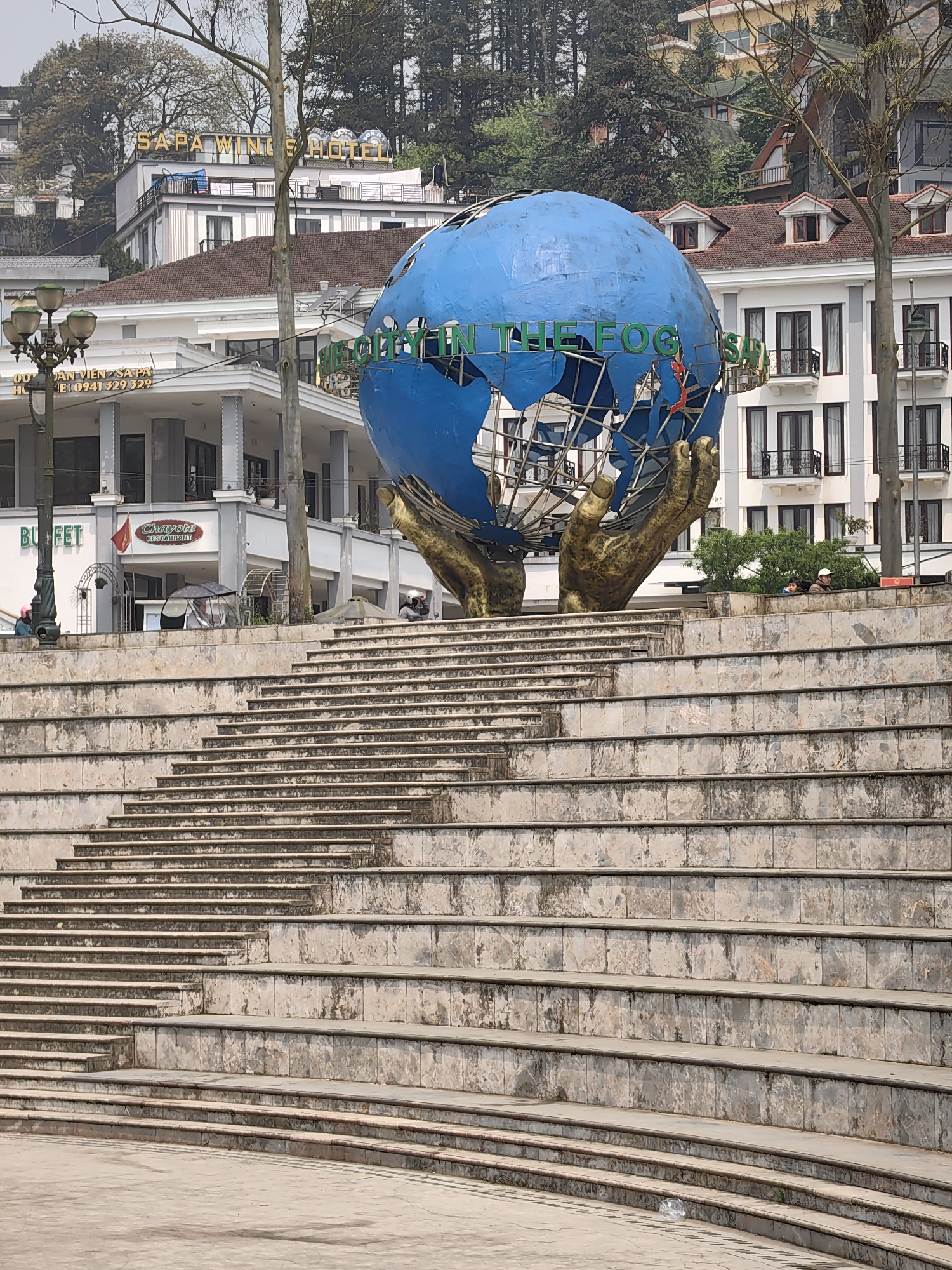 An outdoor wide shot captures a public square in Sa Pa, Vietnam, featuring a large, weathered blue globe sculpture supported by two oversized golden hands. The globe displays partially visible green text, "IN THE FOG" and "SA PA".
In the foreground, a wide, curved amphitheater-style set of stone steps, exhibiting discoloration and grime, descends towards the bottom of the frame. A crumpled clear plastic bottle rests on one of the lower steps.
Behind the steps and the globe, multiple buildings line a hillside. A prominent white multi-story structure with a red-tiled roof and dark-framed windows with small balconies bears a "SAPA WINGS HOTEL" sign at its apex. Adjacent to it, to the left, another white building section with grey pillars features "BUF ET" and "Chayote RESTAURANT" signage, along with a Vietnamese flag.
On an elevated platform behind the steps, near the base of the globe, two individuals are partially visible: one in a dark jacket and light cap, and another to their left in a blue garment. A third individual in a pink hat is partially obscured by an ornate black street lamp on the far left.
Bare tree branches are scattered throughout the upper portion of the frame, and dense green foliage and additional buildings are visible further up the hillside, indicating a mountainous setting under an overcast sky.