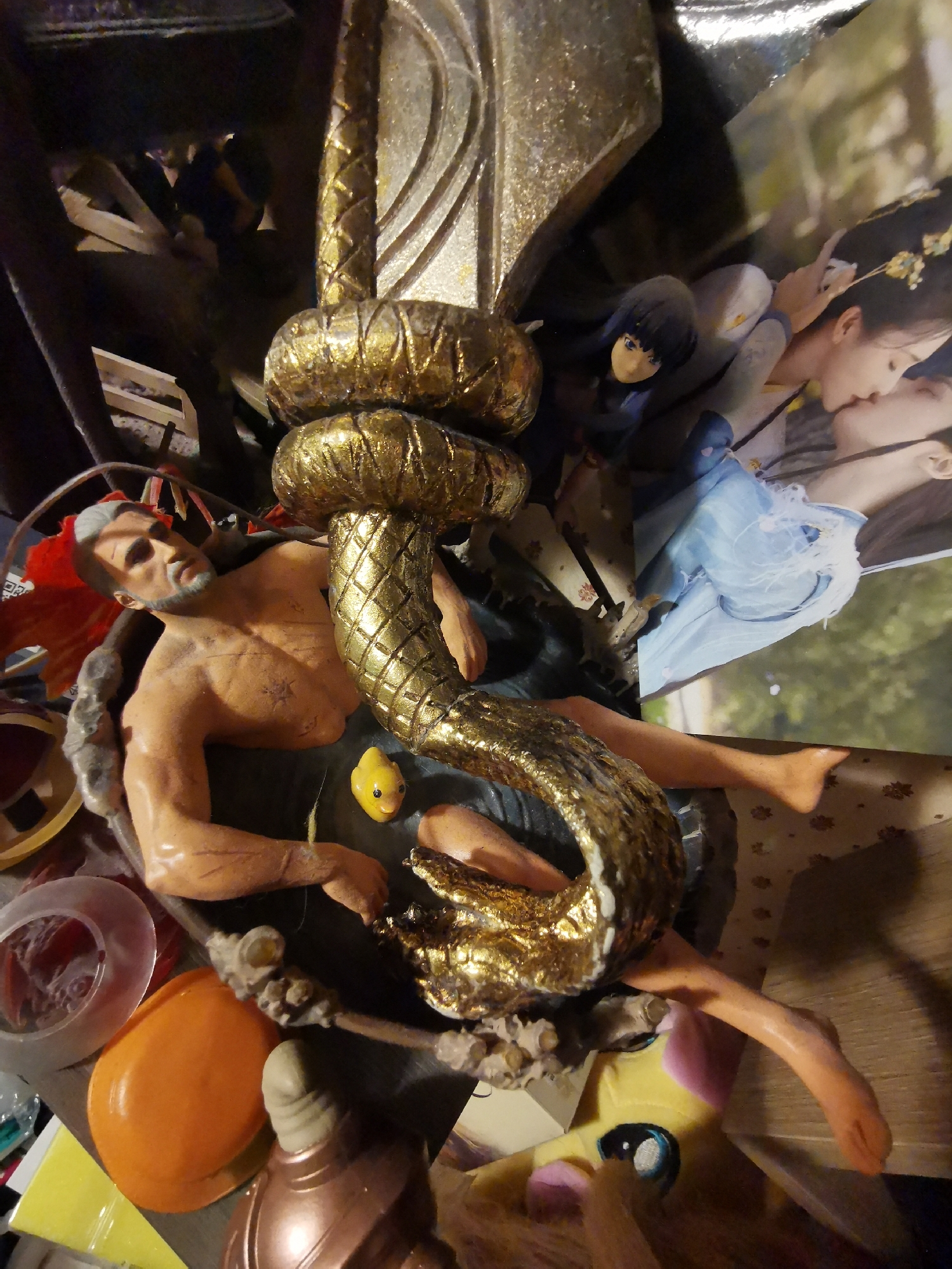 The image depicts a cluttered shelf or table containing various items.
The main subject is a figurine of a muscular man with white hair and a beard, lying in what appears to be a basin or pool. A small yellow rubber duck is next to him. Above him is a gold-colored, twisted metal structure that resembles a staff or ornamental piece.
Behind the figurine is a postcard or photograph depicting two Asian women in a romantic pose. To the side of the photo is a figurine of a girl with black hair and blue eyes.
In the foreground, there are several miscellaneous objects, including clear plastic lids or containers, an orange plastic object (possibly a lid or toy), and the face of a yellow and pink plush toy. Other items are scattered around.
There is nothing in the image to indicate its location in the city of Moscow, Russia.