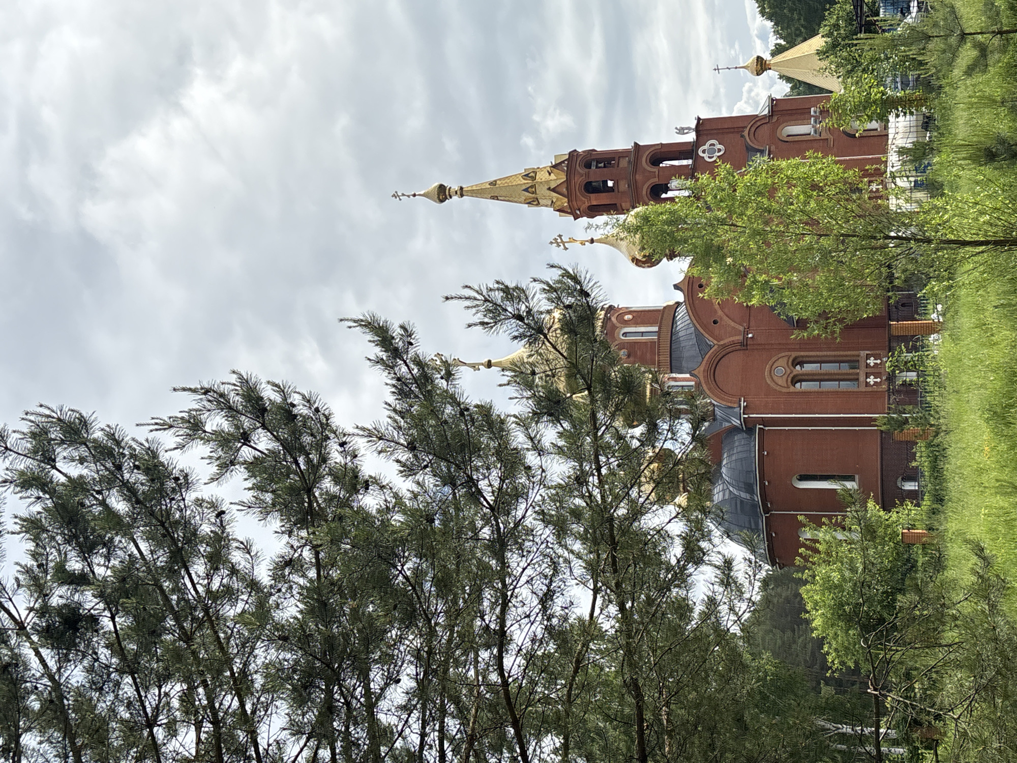 The image displays a majestic Russian Orthodox church, characterized by its red brick construction and brilliant golden onion domes and spires, set against a bright, overcast sky. The building's architecture is intricate, featuring arched windows, decorative elements, and a tall bell tower crowned with a prominent golden spire and a cross. Several other smaller golden domes, each topped with a cross, are visible across the church's roof.
The foreground and midground are filled with dense foliage. On the left, numerous coniferous trees with dark green needles and small cones partially obscure the view, creating a natural frame. Various deciduous trees with vibrant green leaves are scattered throughout the scene, especially on the right, further blending the architectural structure with the natural surroundings. A hint of a fence and possibly other buildings can be seen through the trees on the far right.
The scene is captured during the daytime under a cloudy sky, providing soft, diffused lighting. No individuals are visible. The architectural style, particularly the onion domes, is highly characteristic of Russian Orthodox churches, aligning with the given location of Chaikovsky, Russia. The image evokes a sense of tranquility and spiritual grandeur. There is no discernible text in the media file.