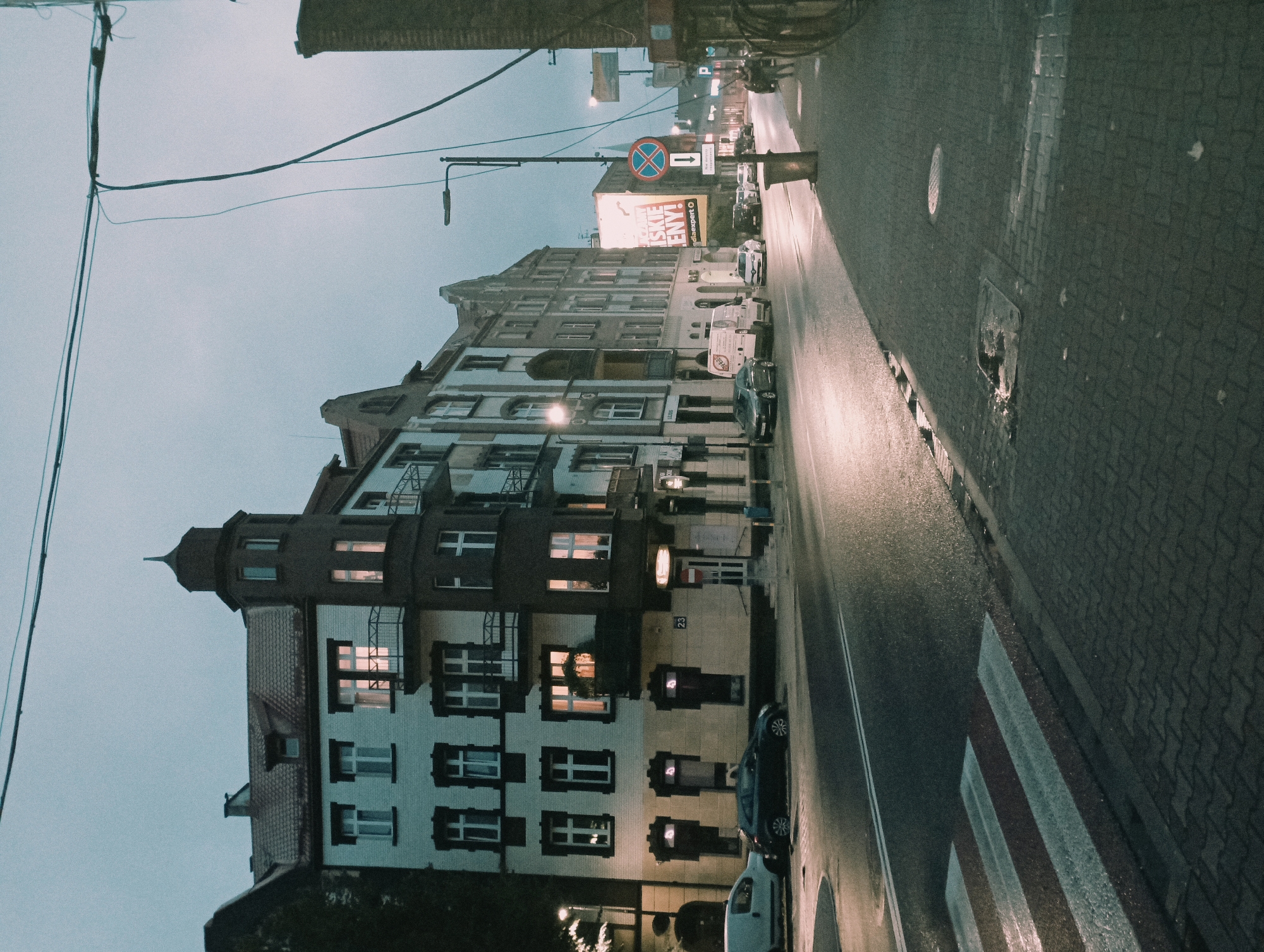 The image depicts a street scene in Siemianowice Śląskie, Poland, characterized by its dim, overcast sky and wet pavement, suggesting recent rainfall. A multi-story building with a distinctive turret dominates the left side of the frame, its façade a mix of white and darker tones. Several windows in the building are illuminated from within. Adjacent to this building, a row of other structures follows the street, some featuring gabled roofs and intricate architectural details. The street itself is asphalt, marked with double white lines, and reflects the artificial light from streetlamps and building interiors. A pedestrian crossing, with alternating white and red stripes, is visible in the foreground, appearing wet and slightly reflective. Parked cars, including a white van and a dark sedan, are present along the roadside. A sidewalk paved with cobblestones runs along the right side of the street, featuring streetlights, signage, and a trash receptacle. In the distance, more buildings and streetlights are visible, contributing to the urban atmosphere. The overall impression is one of a quiet, possibly early morning or late evening, city street.
