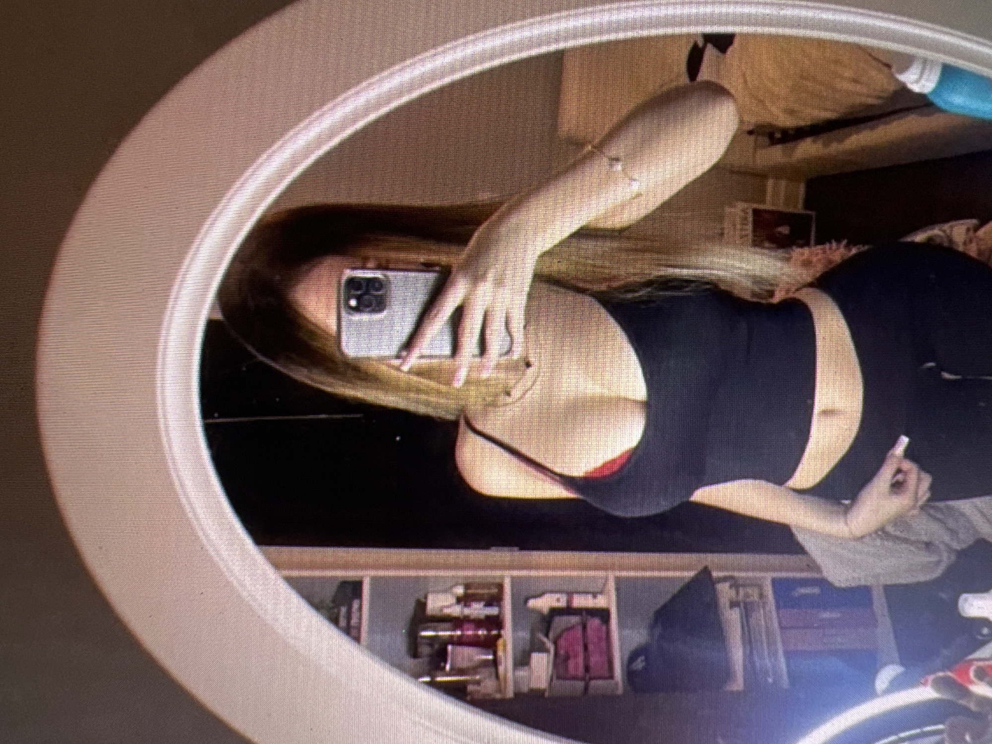 The image shows a young woman taking a mirror selfie in what appears to be a bedroom. She is wearing a black tank top with a red bra underneath and black pants. She has long blonde hair and is holding a smartphone in her right hand to take the picture. In the background, a shelf unit filled with various items, including bottles and boxes, is visible. The room also contains a bed with a tan comforter and a blue object on the floor. A portion of a white oval mirror frame is visible, reflecting the scene. The overall lighting is dim, with some light sources creating reflections on the mirror. There is no information in the image to indicate a specific location in Castelo Branco, Portugal.